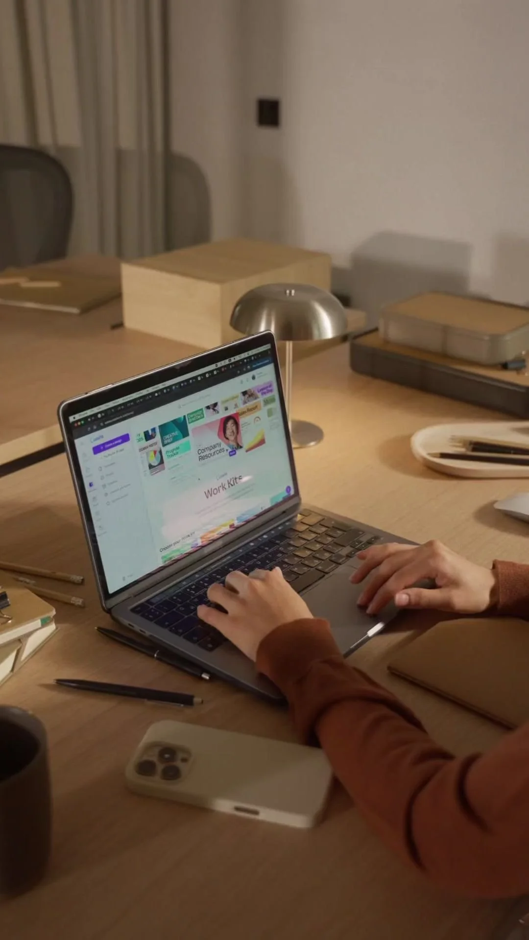 Person working on a laptop at a wooden desk with office supplies, a smartphone, and a desk lamp.