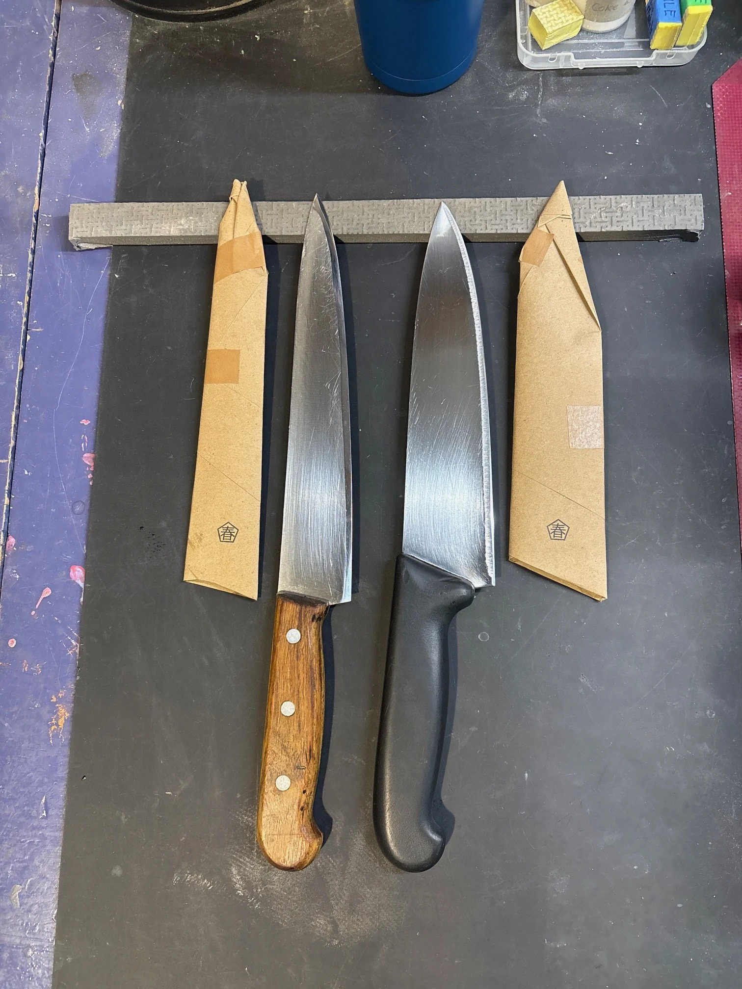 Deux couteaux, l'un avec une lame en acier et manche en bois, et l'autre avec une lame en acier et manche en plastique, reposent sur une surface noire avec deux paquets enveloppés de papier kraft à côté.