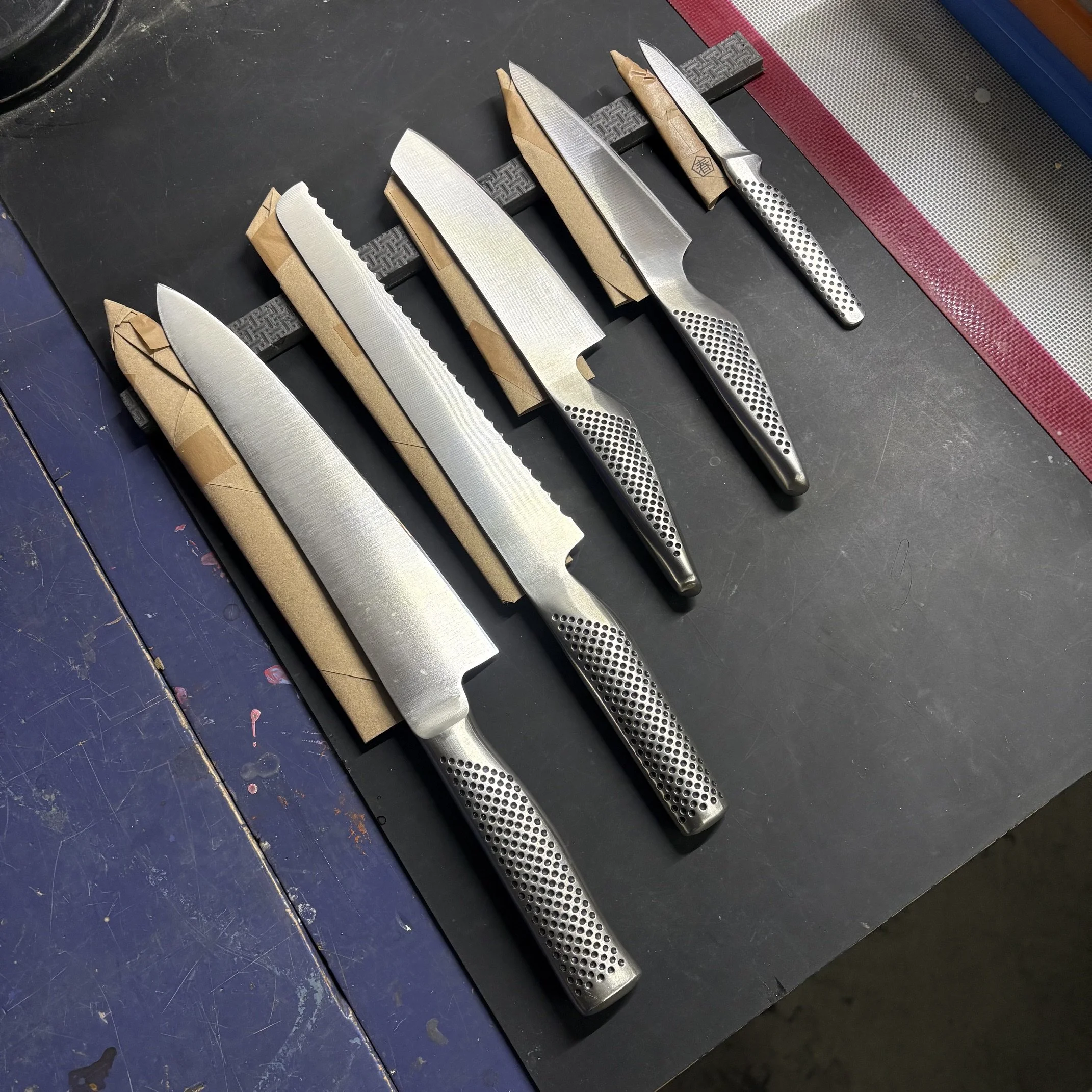 Cinq couteaux en acier avec des manches en métal perforé ou en papier craft, posés sur un support en plastique noir, sur une surface en bois peint, avec un tapis en silicone ou en caoutchouc à droite.