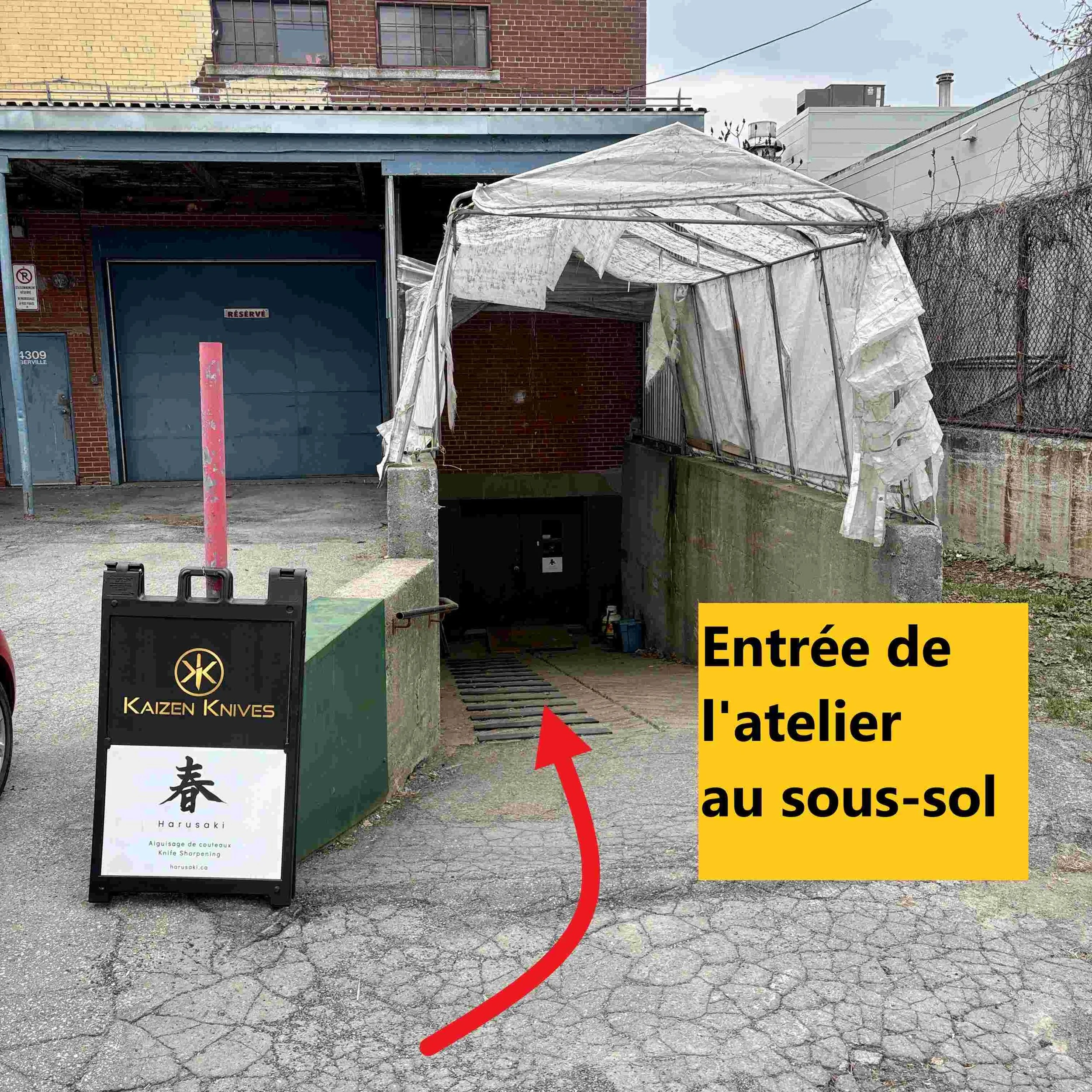 Un panneau indicateur jaune avec du texte en français indiquant l'entrée de l'atelier sous-sol, une façade de bâtiment en briques rouges avec une porte bleue réservé, une tente blanche partiellement déchirée, un panneau publicitaire à gauche avec des caractères japonais et le nom Harusaki, une clôture en métal, un arbre sans feuilles et un sol en pierre craquelée.