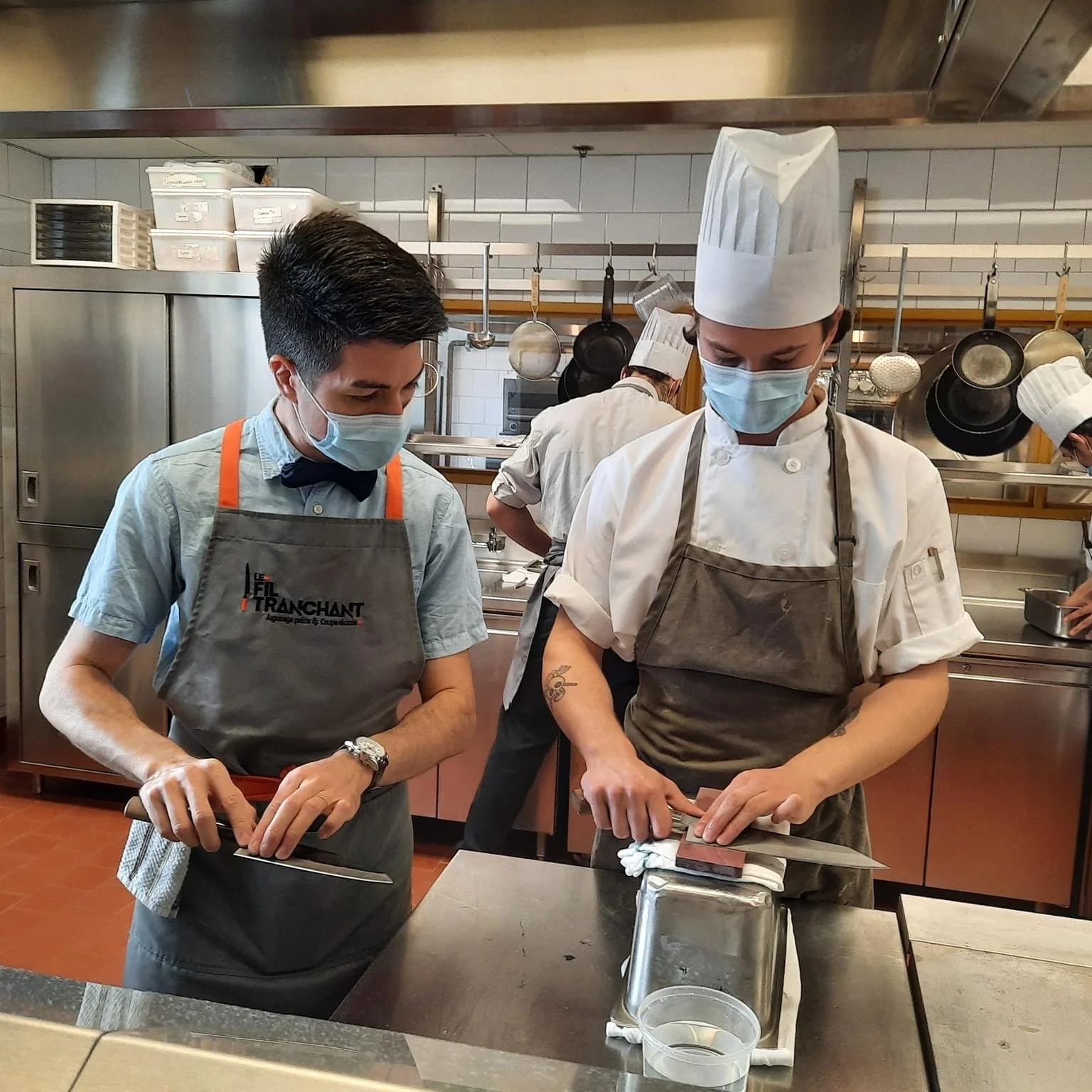 Un instructeur et un chef dans une cuisine, tous les deux portant un masque hygiénique. L'instructeur à gauche est en train de démontrer le mouvement et le chef aiguiser son couteau sur une pierre d'aiguisage à l'eau.