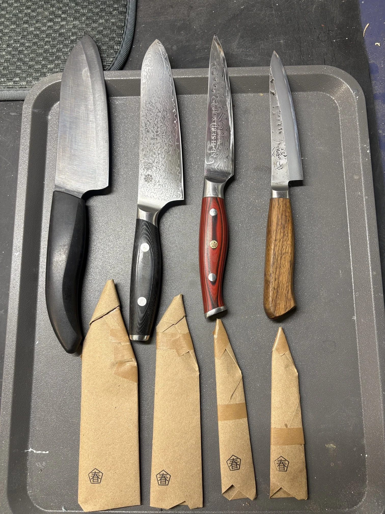 Couteaux de cuisine posés sur une plaque en métal, certains avec des poignées en bois ou en matériau noir, rangés avec des étuis en papier kraft en dessous.