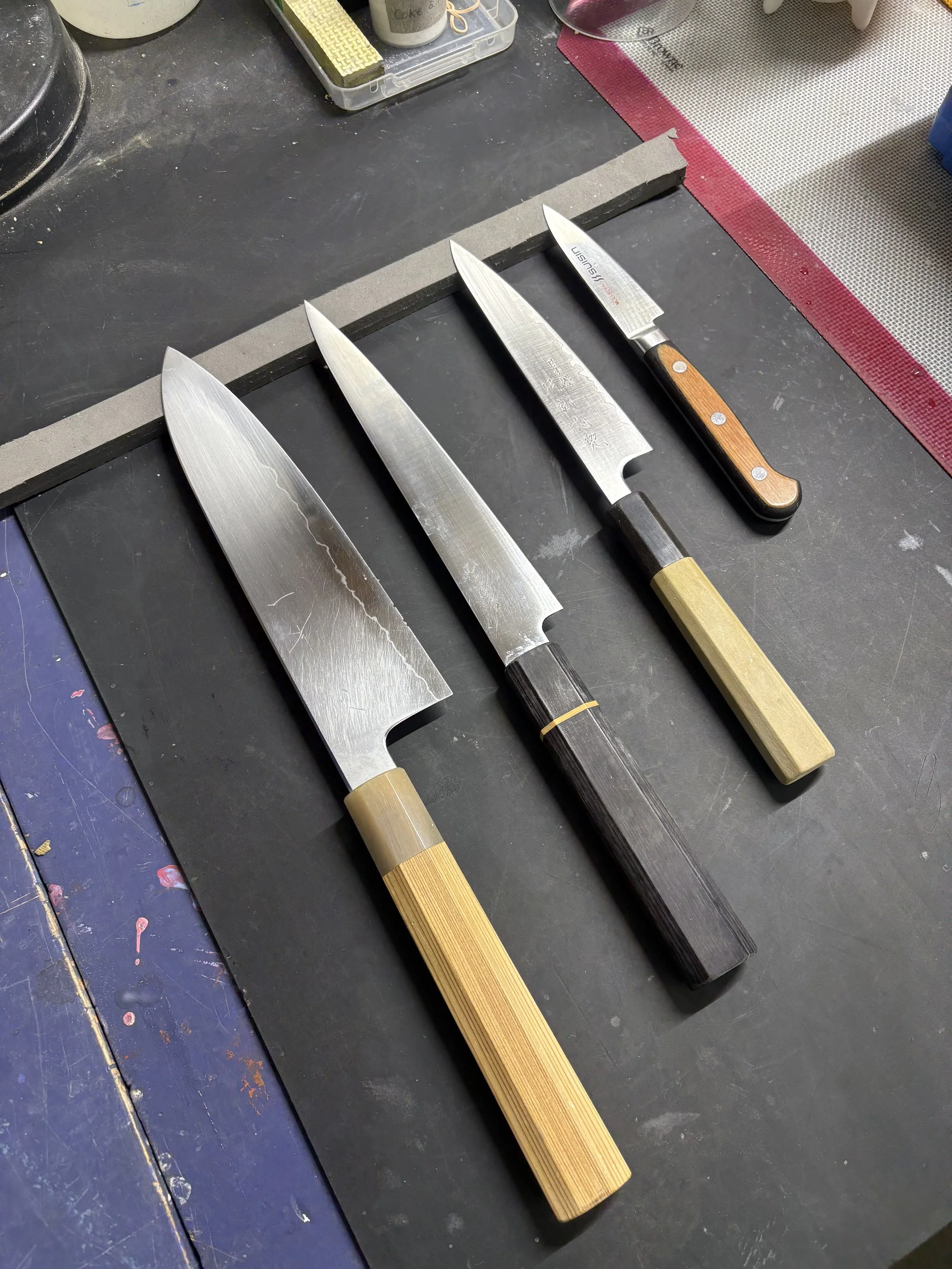 Quatre couteaux de cuisine avec différentes lames et manches en bois, posés sur un plan de travail noir, avec des ustensiles et un tapis de découpe en arrière-plan.