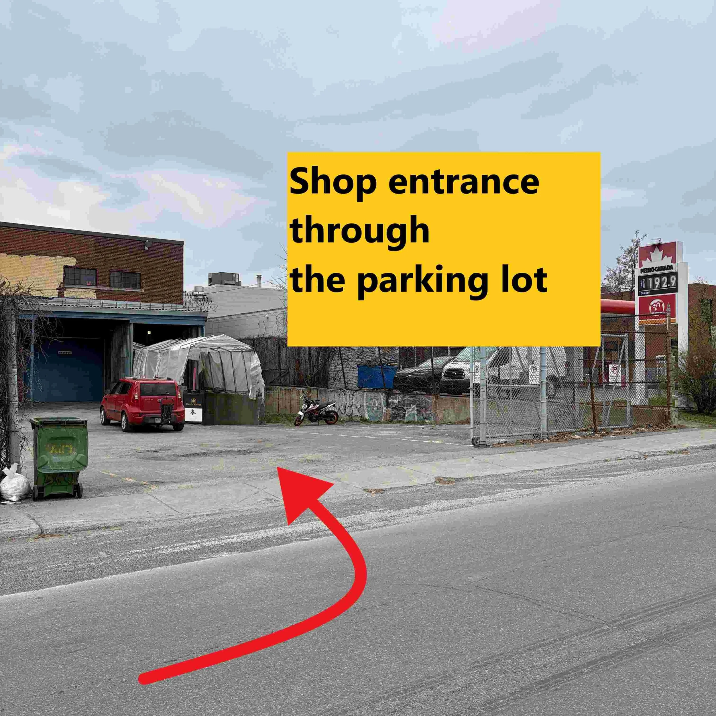 Un panneau jaune indique l'entrée du magasin, avec une flèche rouge pointant en direction du sol devant un parking extérieur avec des voitures, des motos et une zone clôturée.