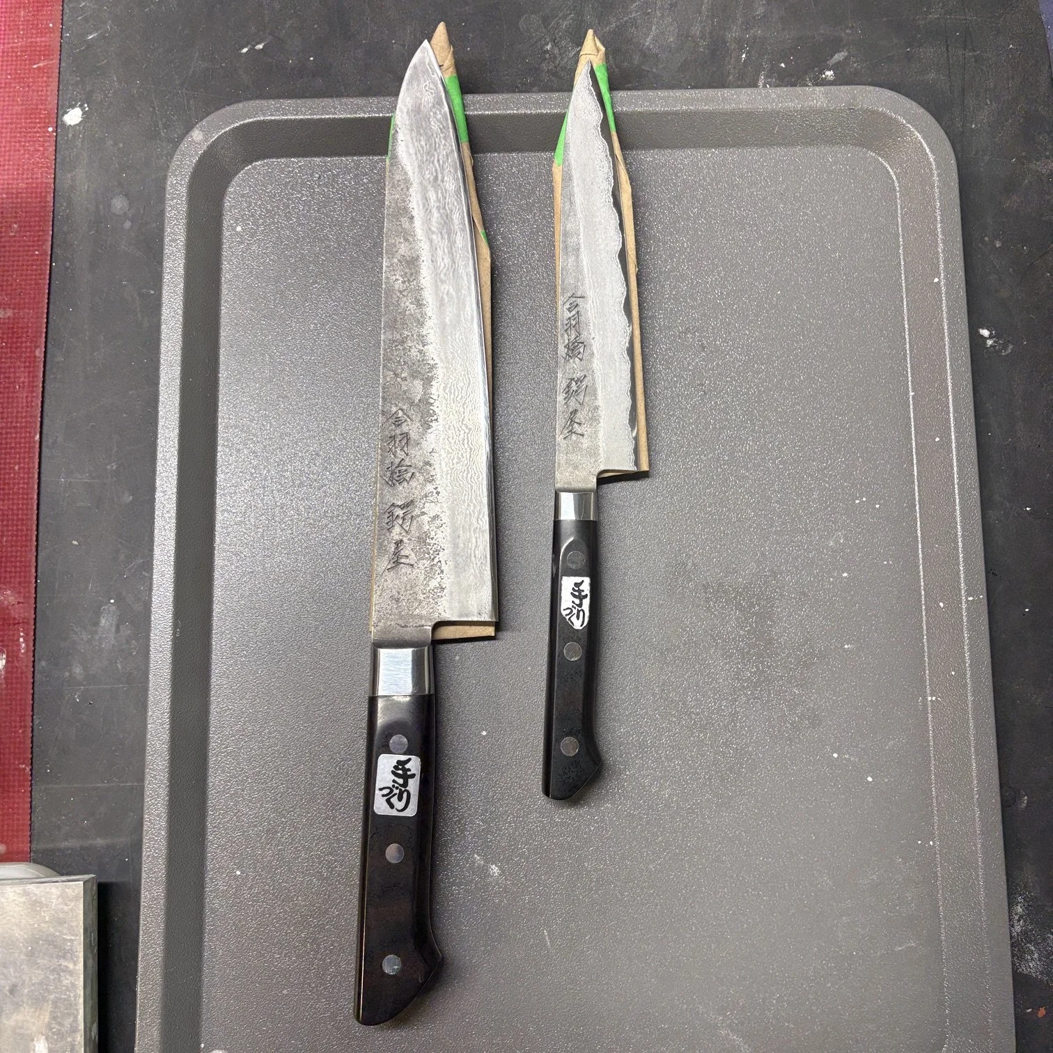 Deux couteaux de cuisine japonais avec des lames en acier inoxydable et des manches en bois, disposés sur un plateau en plastique gris.