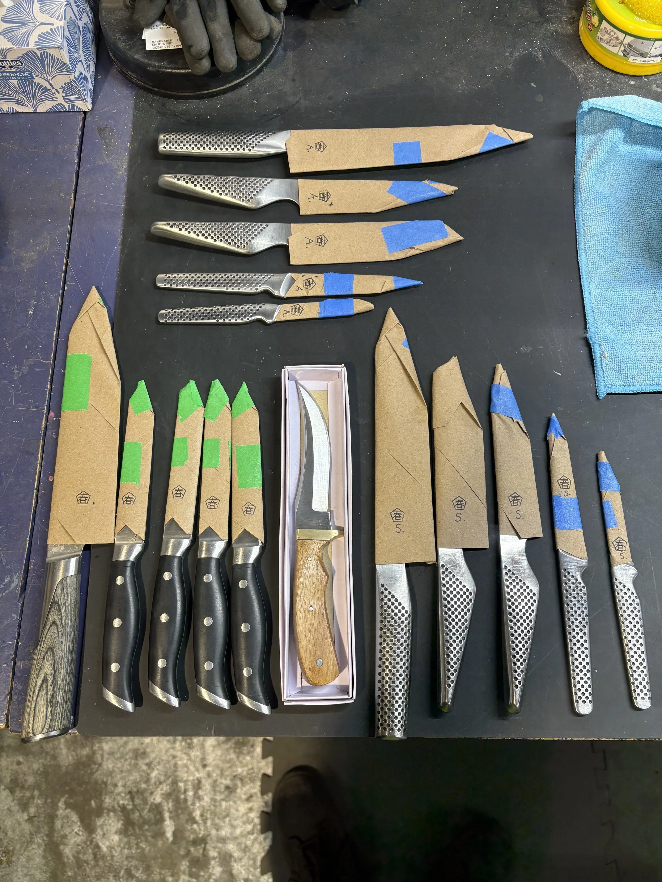 Set de couteaux de cuisine avec manches en métal et en bois, certains dans des enveloppes en papier avec des marques de protection en ruban bleu, noir. À côté, une boîte rectangulaire blanche et un tapis en tissu bleu. Quelques outils ou matériaux di