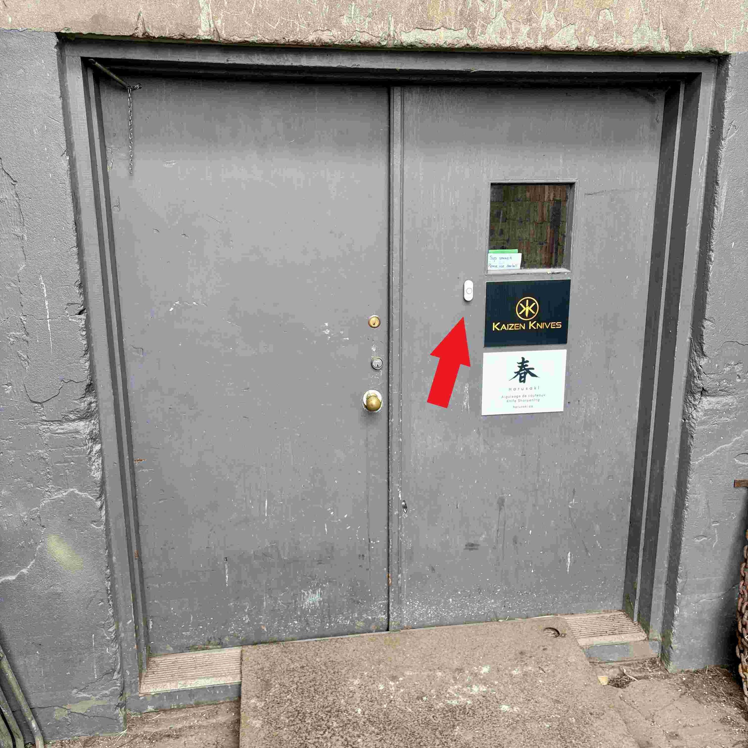 Porte en métal grise avec panneau et petite fenêtre, entourée d’un mur en béton, avec un panneau indiquant 'Kaizen Knives'