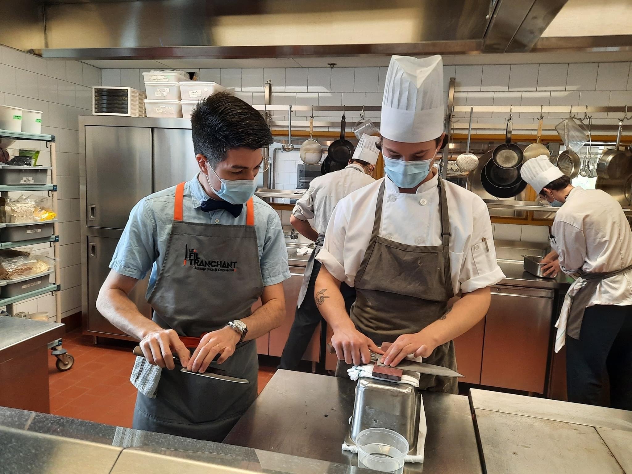 Un instructeur et un chef dans une cuisine, tous les deux portant un masque hygiénique. L'instructeur à gauche est en train de démontrer le mouvement et le chef aiguiser son couteau sur une pierre d'aiguisage à l'eau.