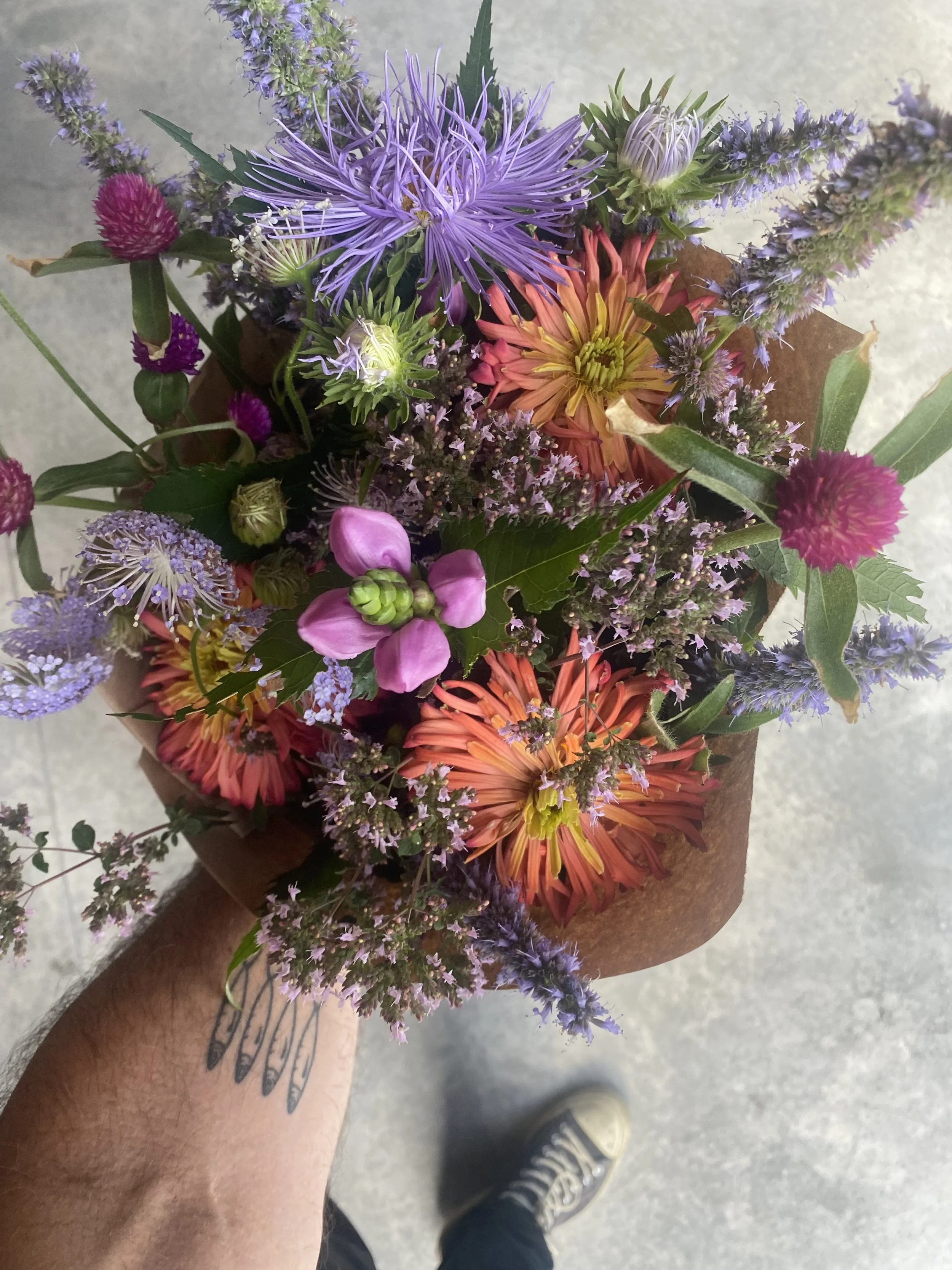 Un bouquet de fleurs colorées comprenant des chrysanthèmes, des lavandes, des œillets et d'autres petites fleurs, enroulé dans du papier craft, vu du dessus.