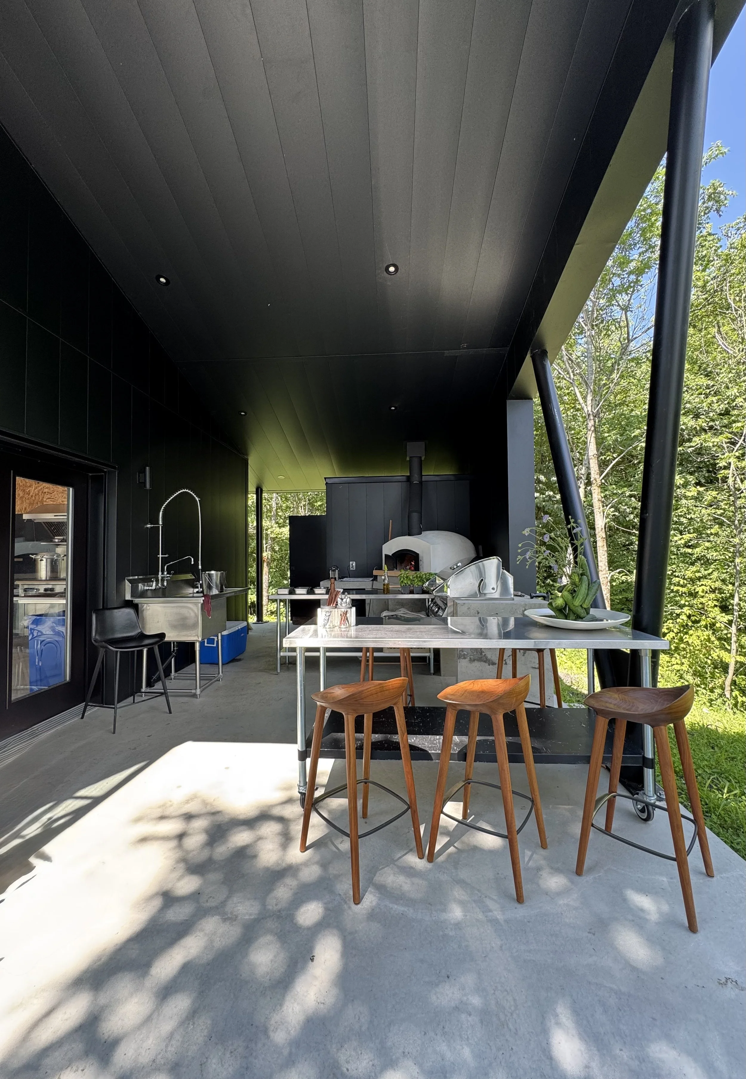 Une cuisine extérieure moderne avec un bar en bois, trois chaises en bois, un four à pizza en pierre, une table en acier inoxydable, un évier, et un environnement verdoyant.