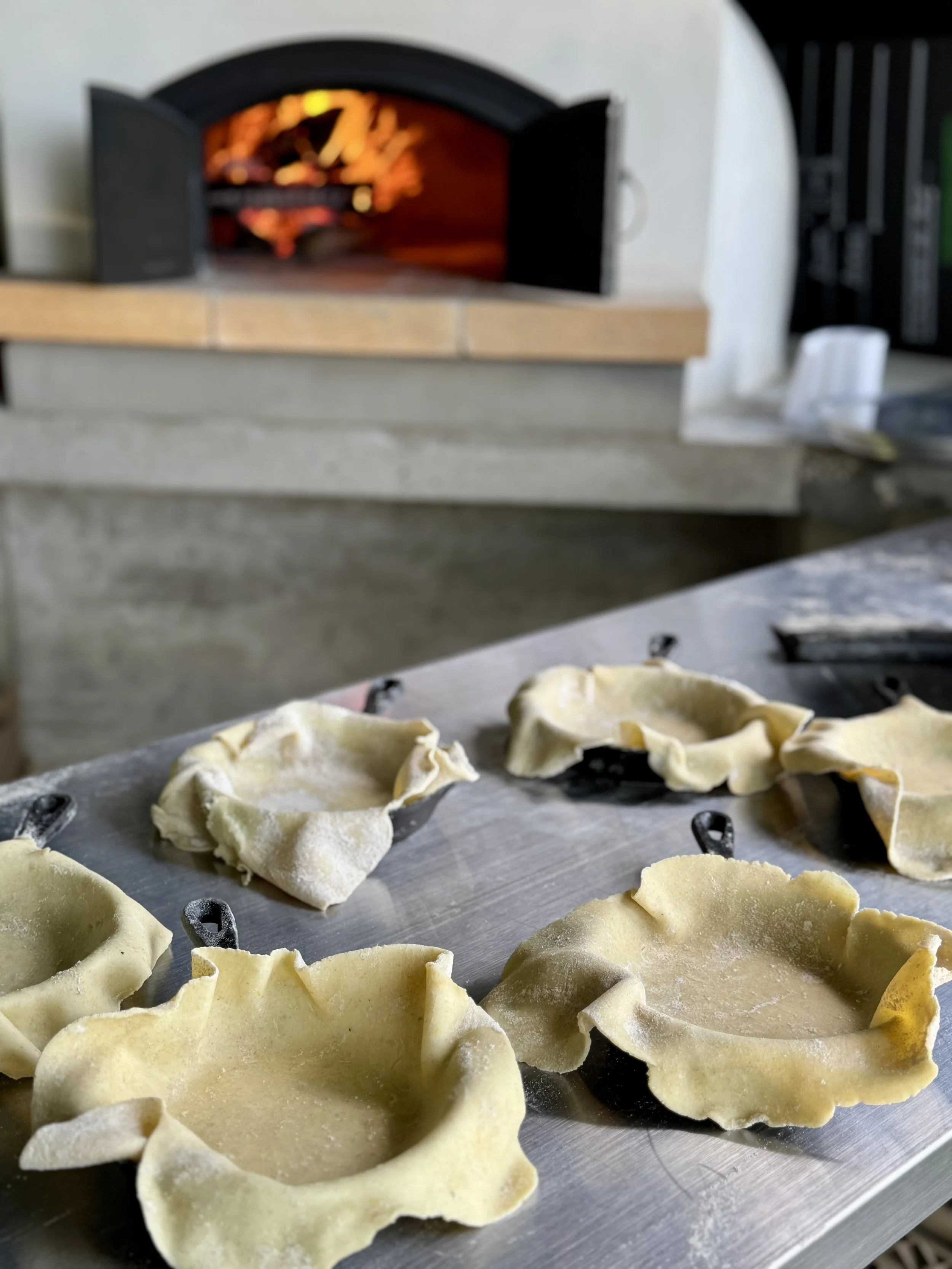 Des mini-tartes en préparation sur une plaque en métal, avec un four à bois en arrière-plan.