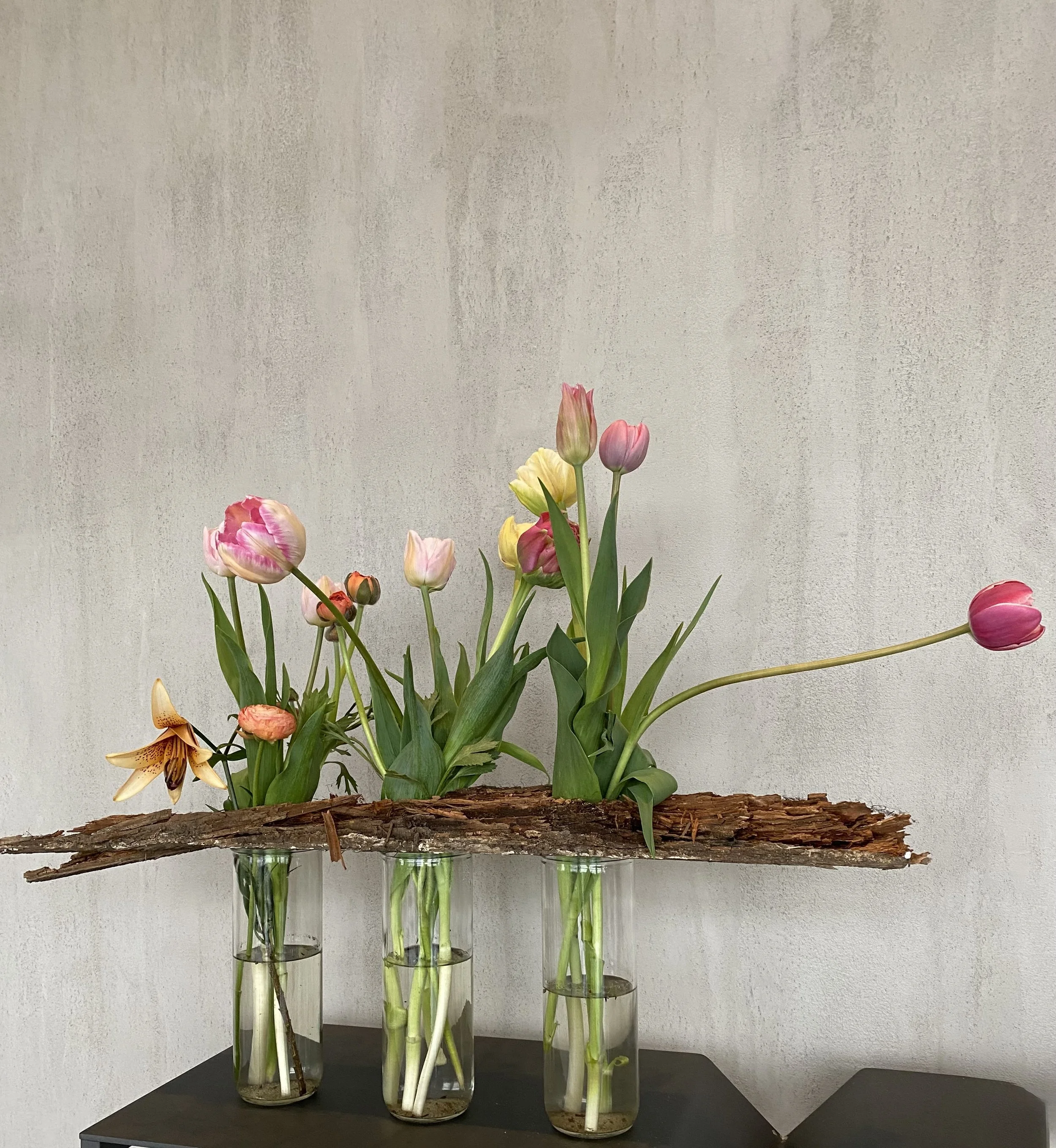 Arrangement floral avec tulipes et autres fleurs dans des vases en verre posés sur une surface noire. Un morceau de bois est placé horizontalement au-dessus des vases.