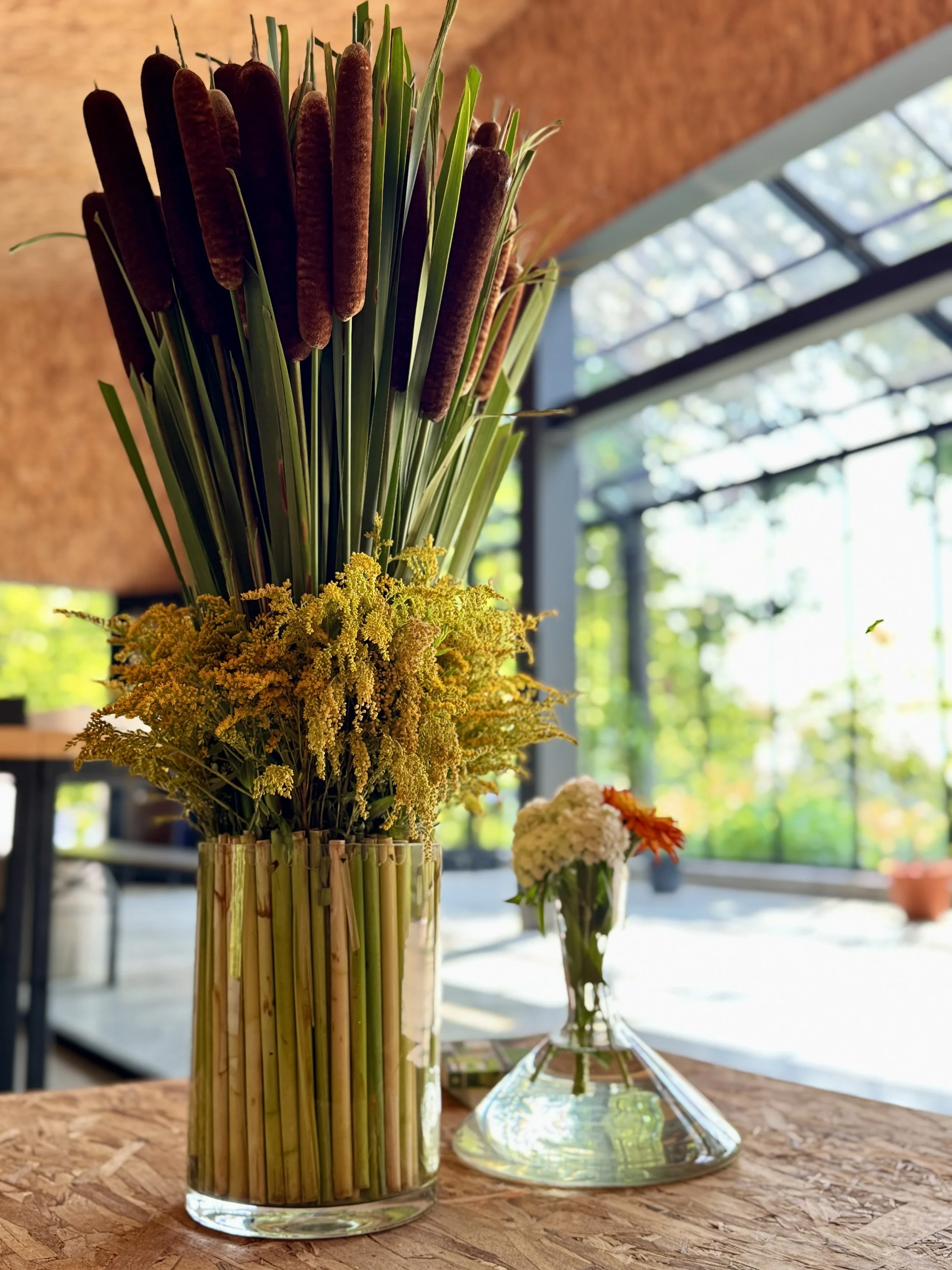 Vase avec un bouquet de fleurs jaunes et vertes, comprenant des végétaux longues et fines, et une petite vase avec des fleurs blanches et rouges, placé sur une table en bois dans une pièce lumineuse avec grande fenêtre.