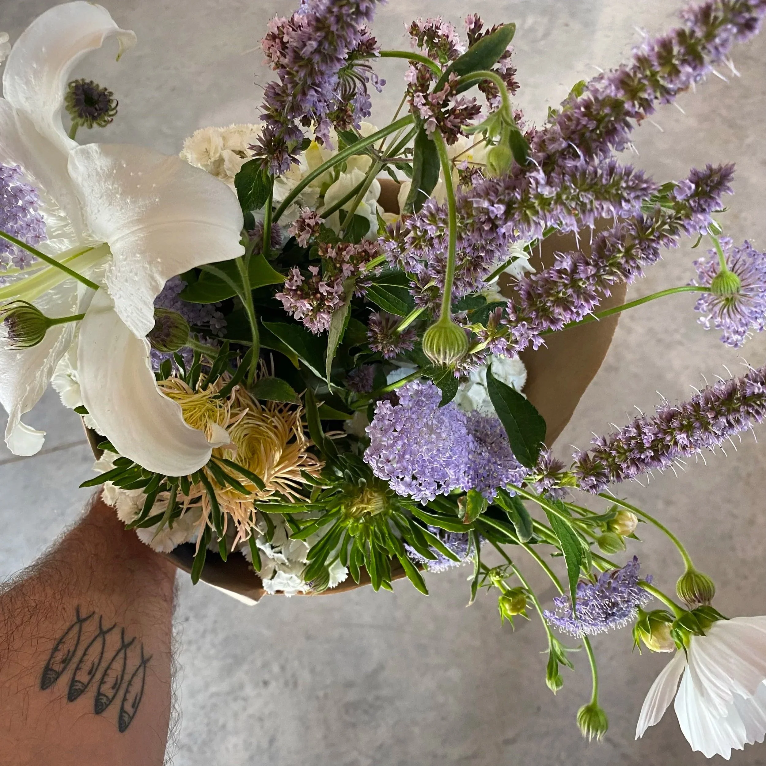 Arrangement floral avec lys blancs, hortensias violets, lavatères violettes, et autres fleurs dans un vase en carton.
