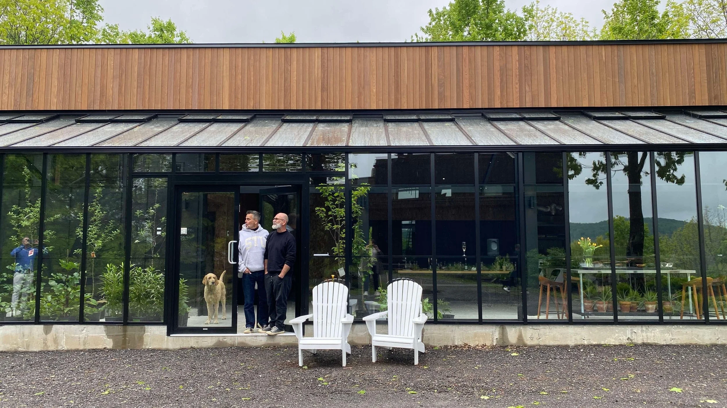 Deux hommes avec un chien à l'entrée d'une serre en verre.  Le bâtiment principal de la micro-ferme maraîchère "Les voisins & Cie"