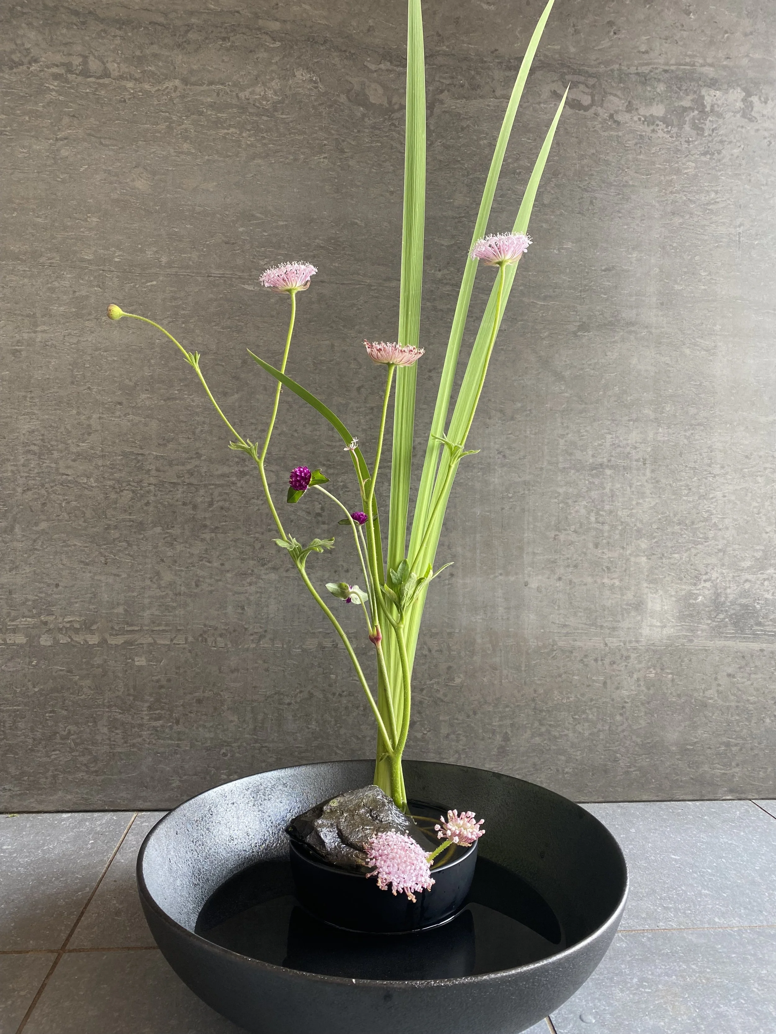 Une composition floral minimaliste avec des fleurs de couleurs roses et violet dans un vase noir posé dans un grand plat noir, contre un mur en pierre grise.