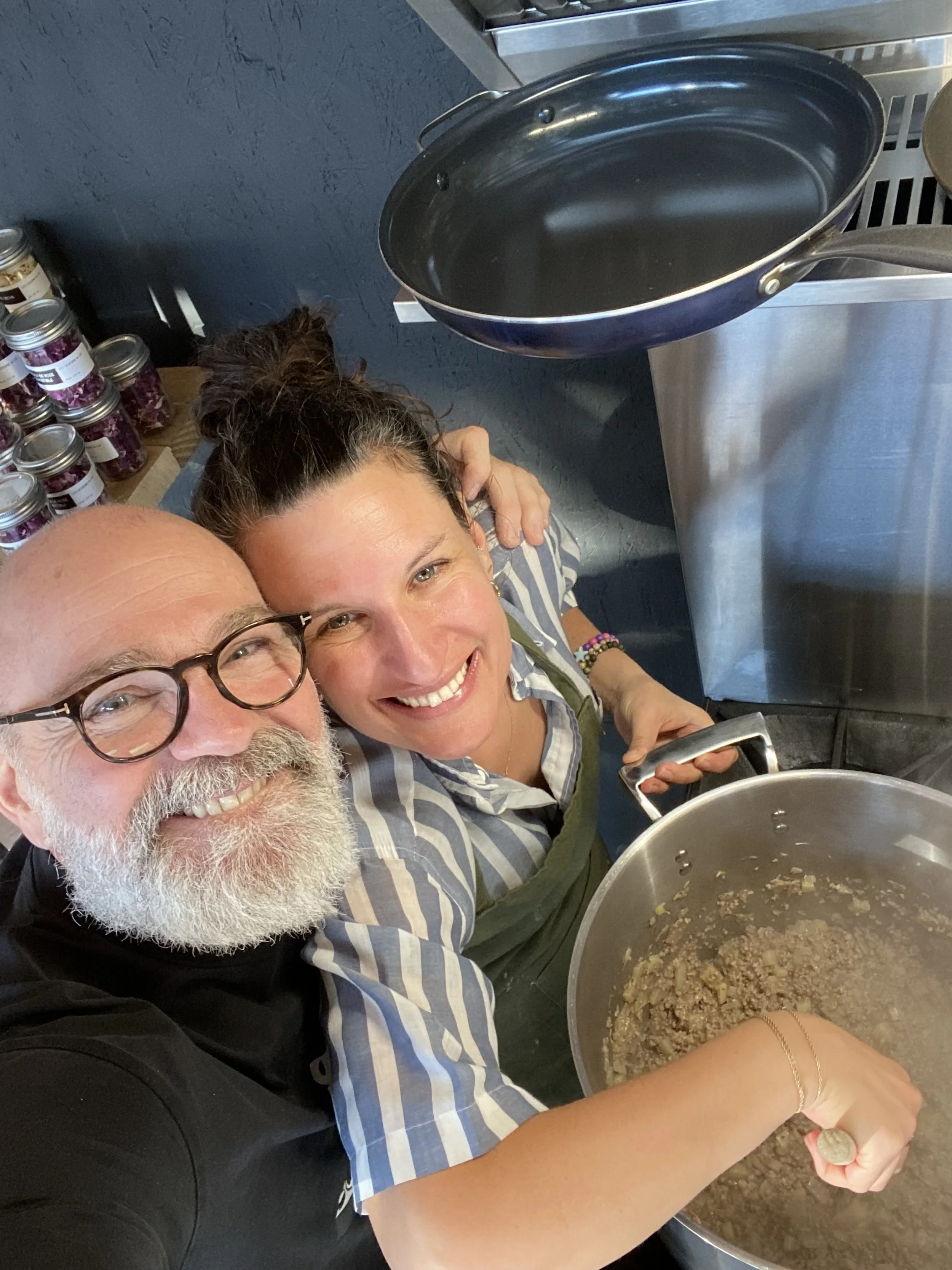 Deux personnes souriantes prennent un selfie dans une cuisine. L'une d'elles remue une grande casserole avec de la viande ou un ragoût, l'autre tient un smartphone. Il y a une surface de comptoir derrière avec des bocaux en verre.