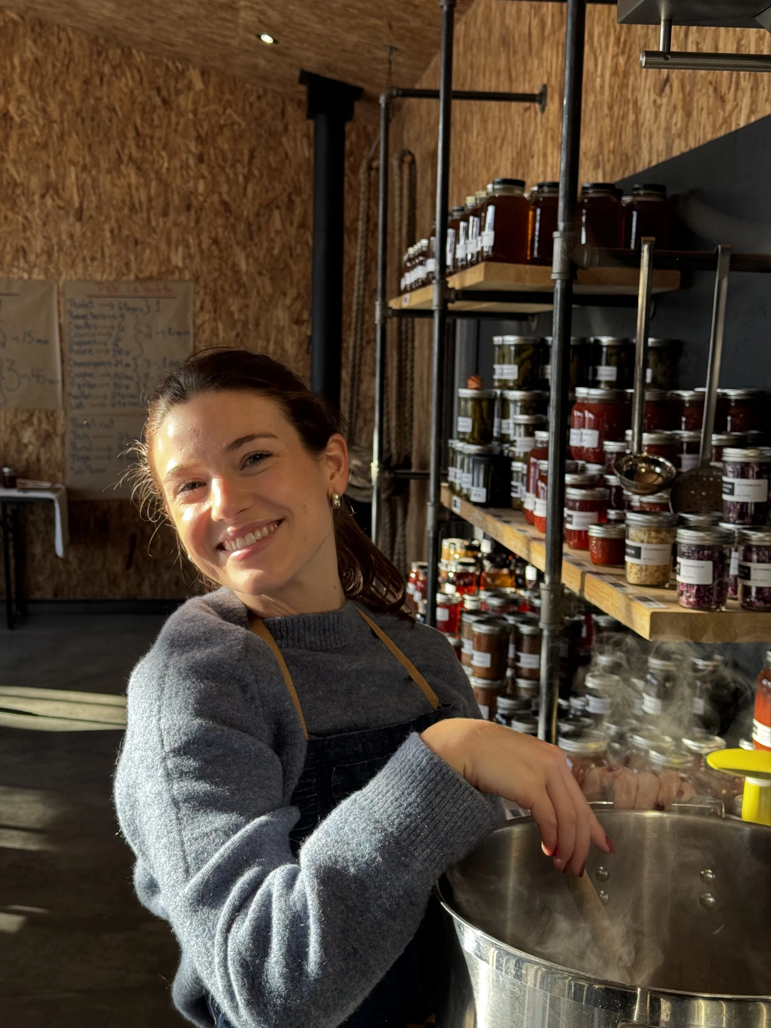 Femme souriante en cuisine, préparant quelque chose dans une grande casserole, avec étagères de bocaux derrière elle.