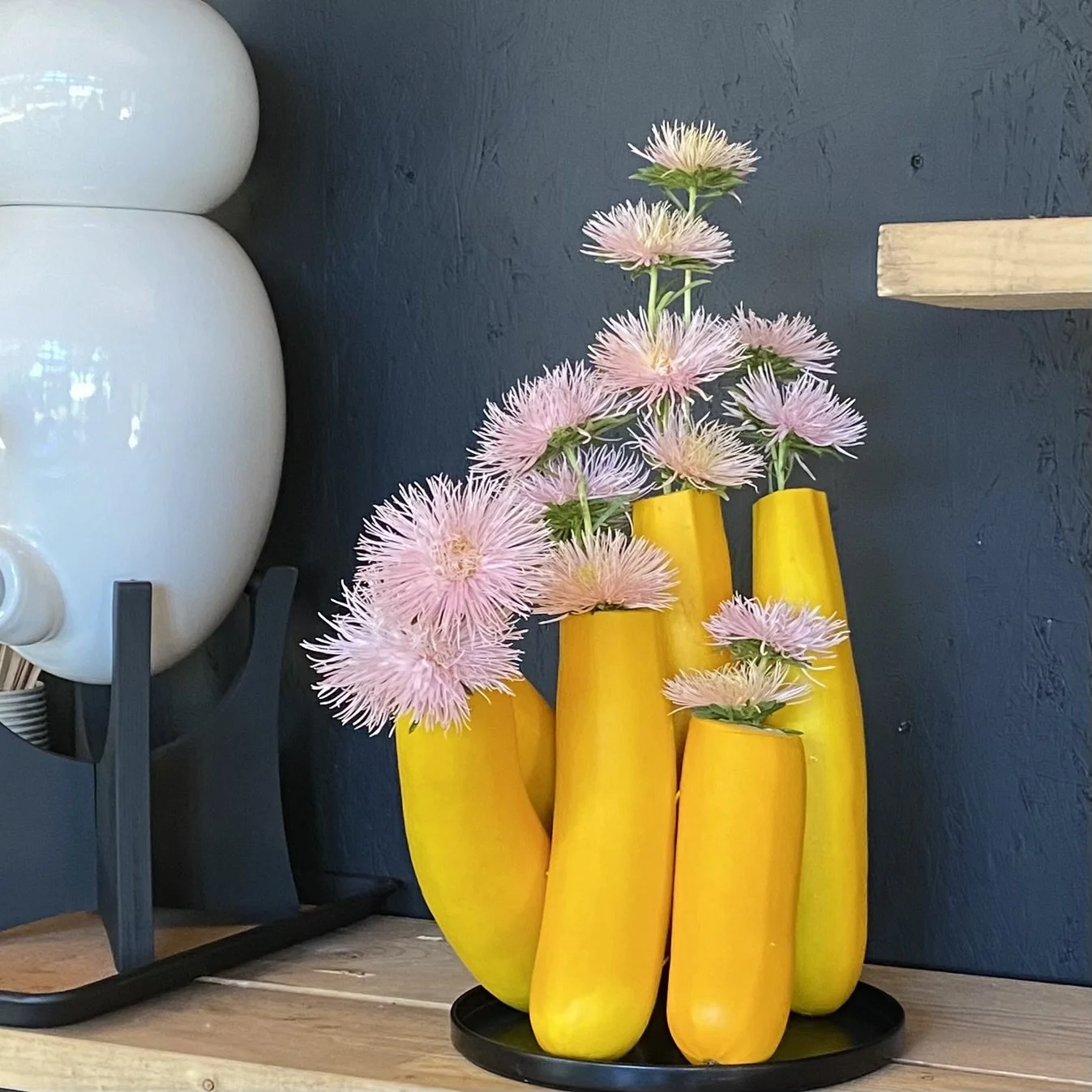 Vase décoratif jaune en forme de bananes avec des fleurs roses et blanches, placé sur une surface en bois, contre un mur gris foncé.