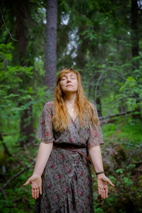 A woman standing in the forest with her hands out and her eyes closed.