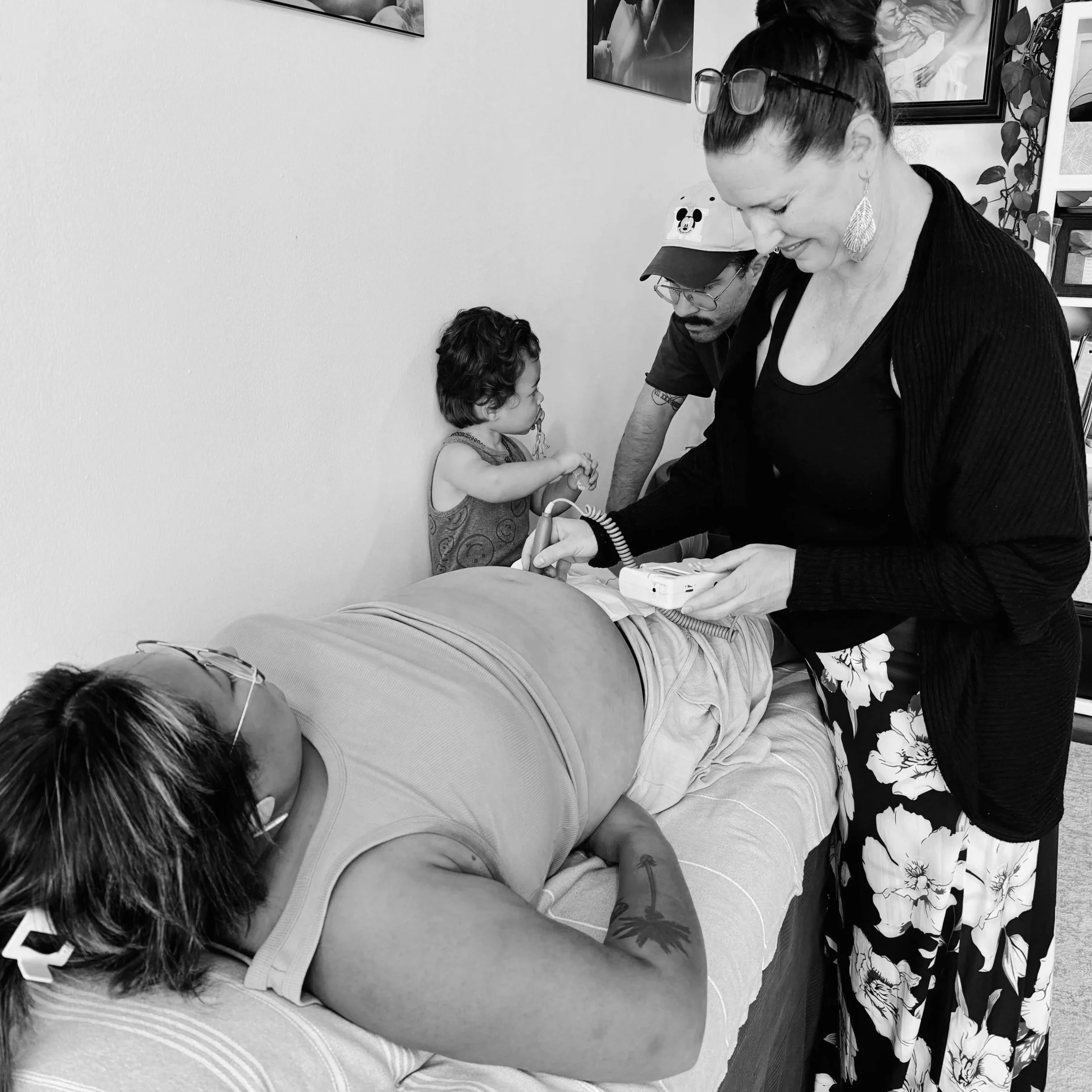 A pregnant woman lying on her back while her midwife listens to baby's heart tones using a fetal doppler, surrounded by her child and husband.