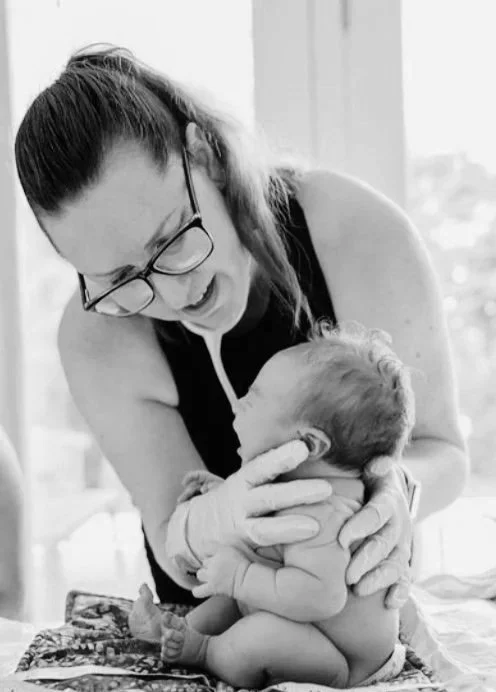 A midwife with her hair tied back wearing glasses and smiling is holding a newborn baby, and examining the baby in the comfort of the family's home. 
