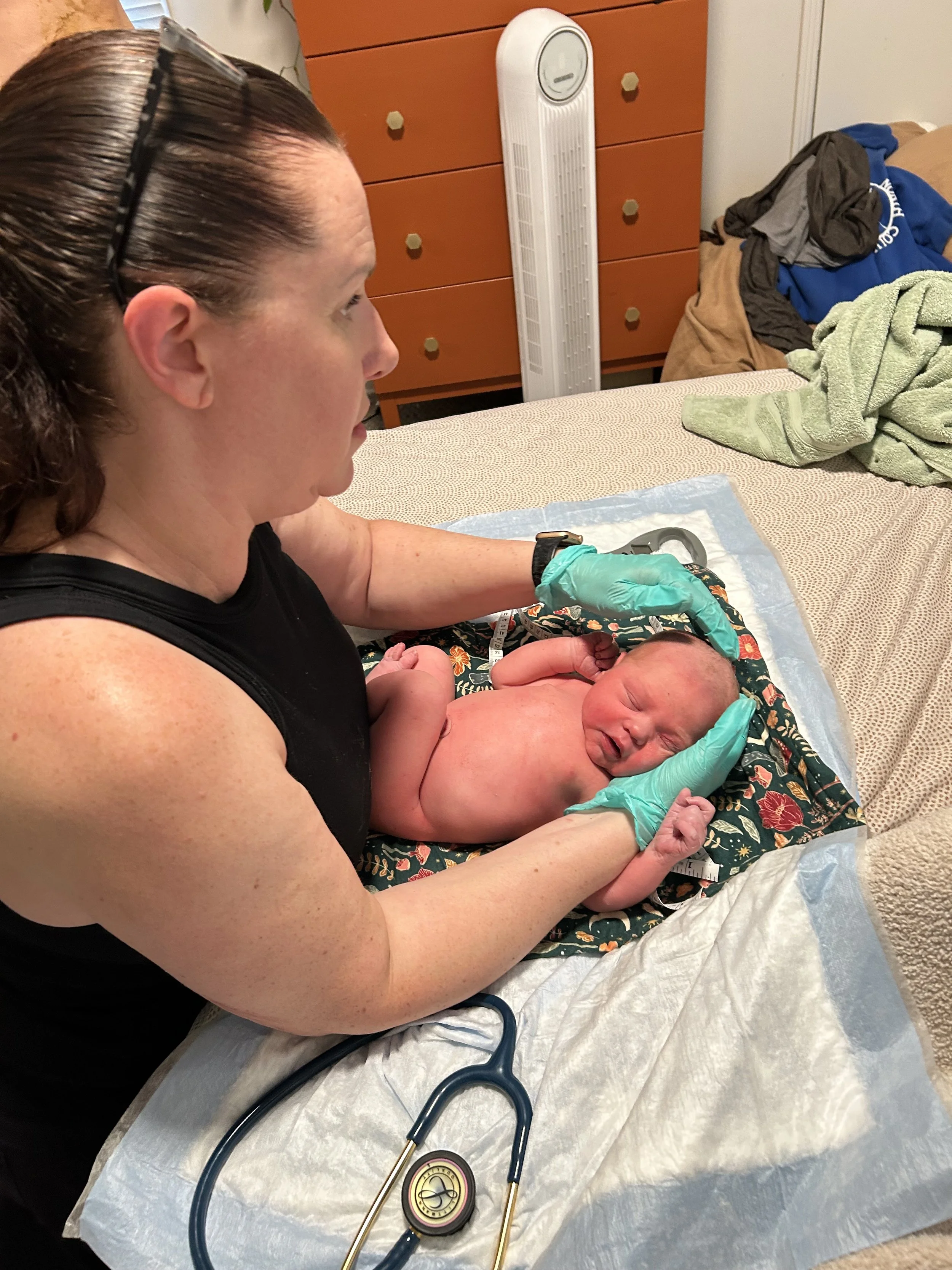 A midwife in black shirt and teal gloves holding a newborn baby performing a newborn exam. The newborn is lying on a floral blanket. Medical equipment, including a stethoscope, is visible on the bed.
