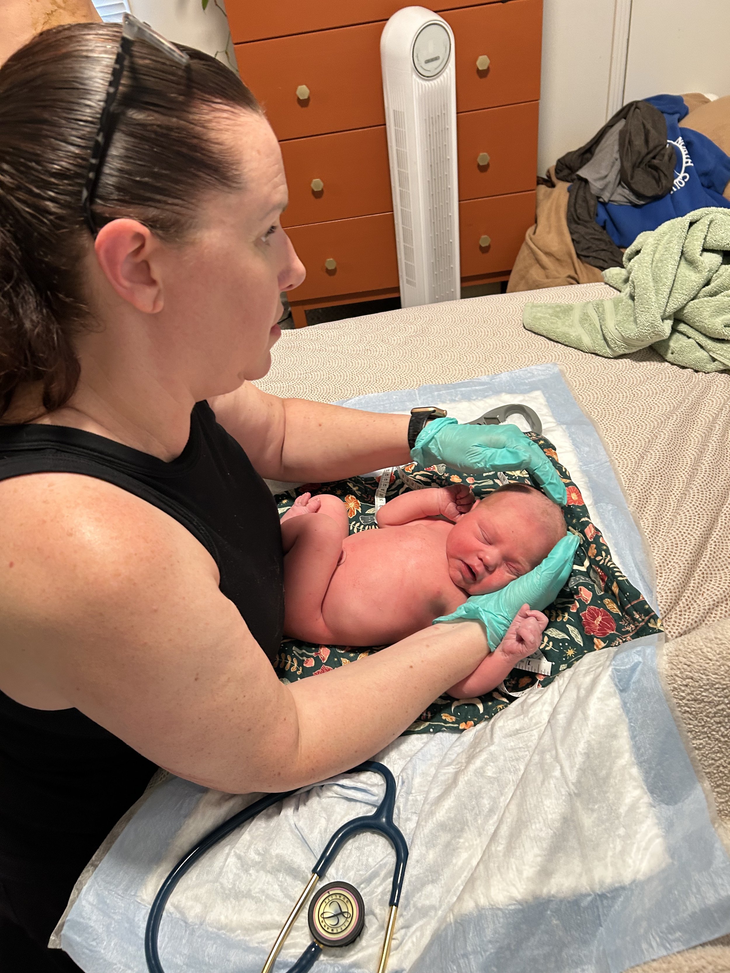 A midwife wearing gloves is holding and examining a newborn baby . The baby is lying on a floral blanket, and the woman has a stethoscope nearby. There is a fan, a dresser, and some clothes in the background.