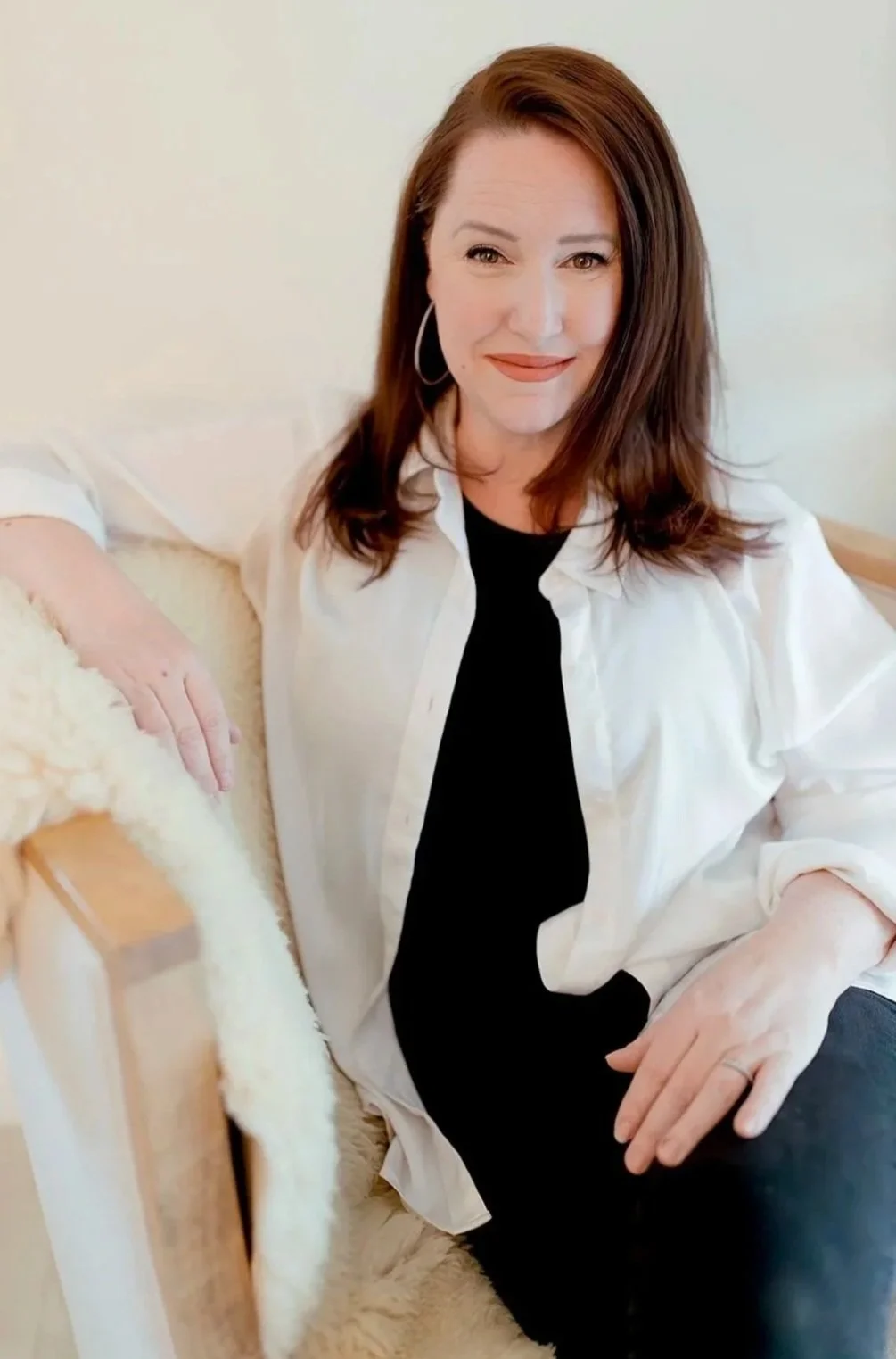 Los Angeles based Midwife Jennifer Buonamassa is posing for a photograph with brown hair,  hoop earrings sitting on a beige couch, wearing a white shirt over a black top, smiling at the camera.
