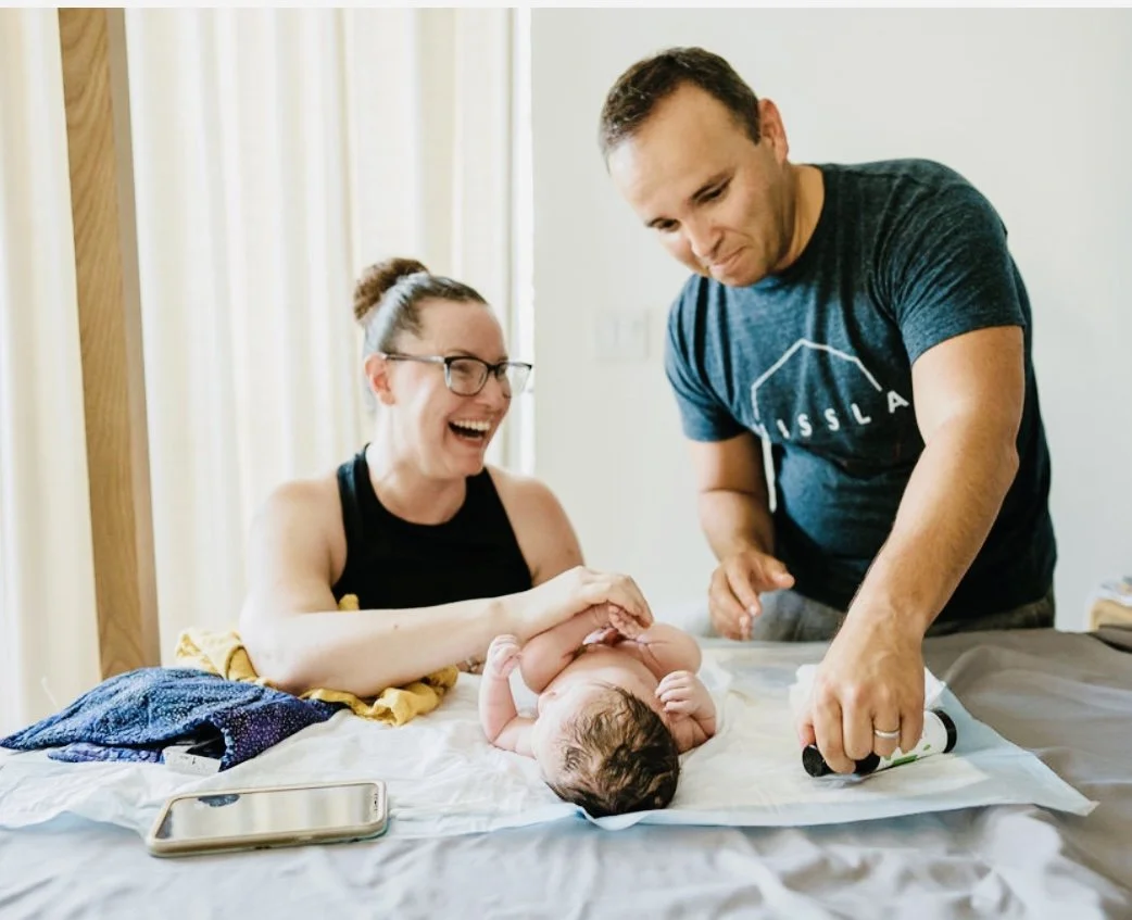 A midwife doing a newborn exam with the baby's father helping, both smiling and laughing as they examine and measure the newborn baby on a bed in a bright room, with a smartphone lying nearby.
