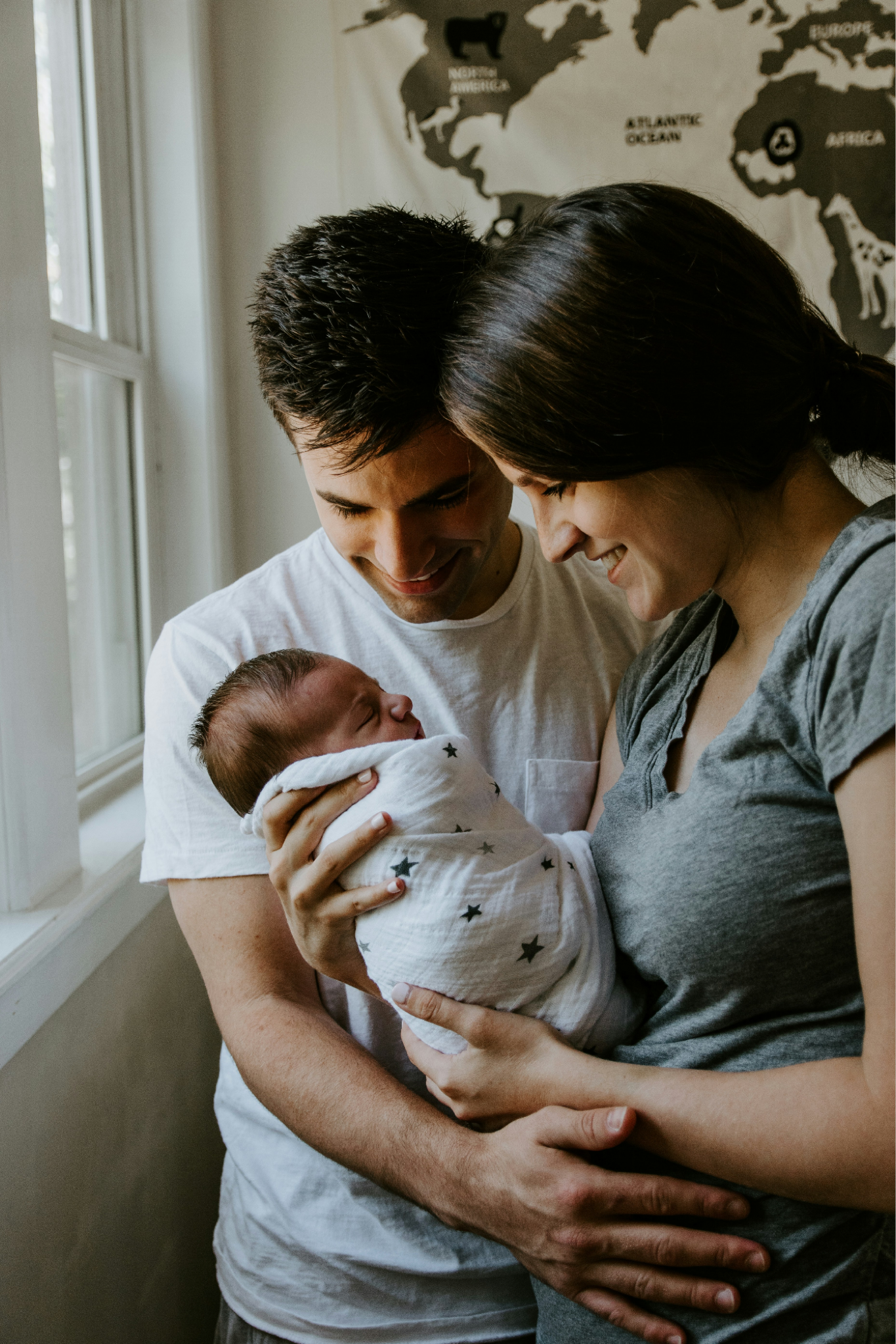 A young couple holding their newborn baby near a window, smiling and looking lovingly at the child.