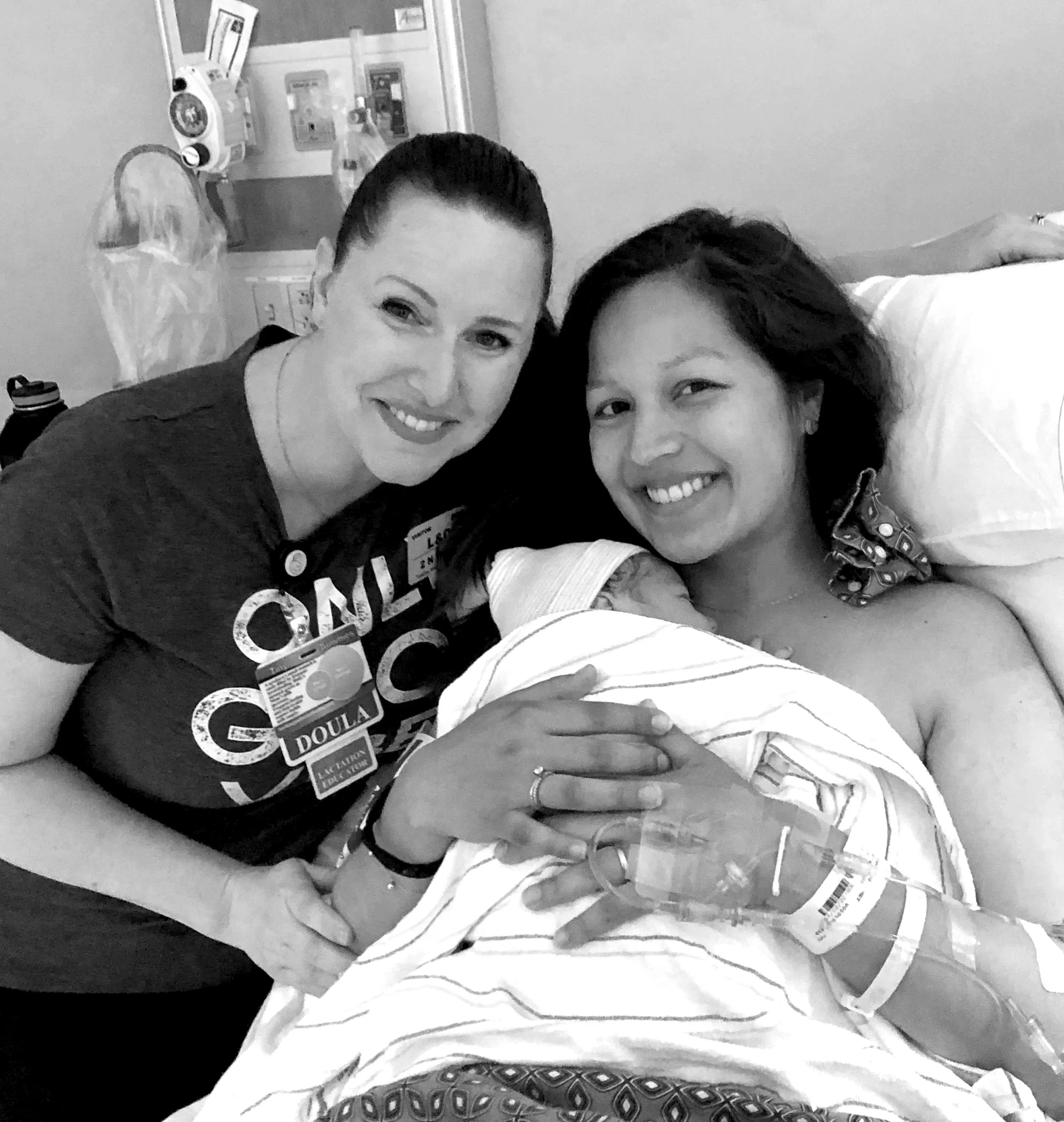 Two women, one in a hospital gown who just gave birth and is holding her newborn baby in a hospital bed, is next to her doula.