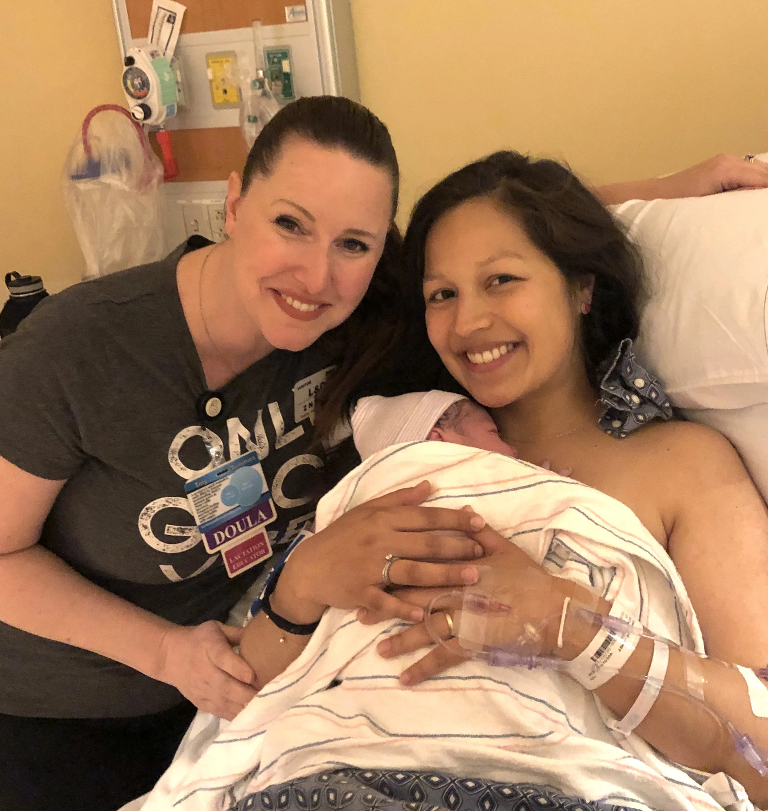 A woman in a hospital bed holding a newborn baby wrapped in a hospital blanket, smiling while her doula stands next to her, also smiling.