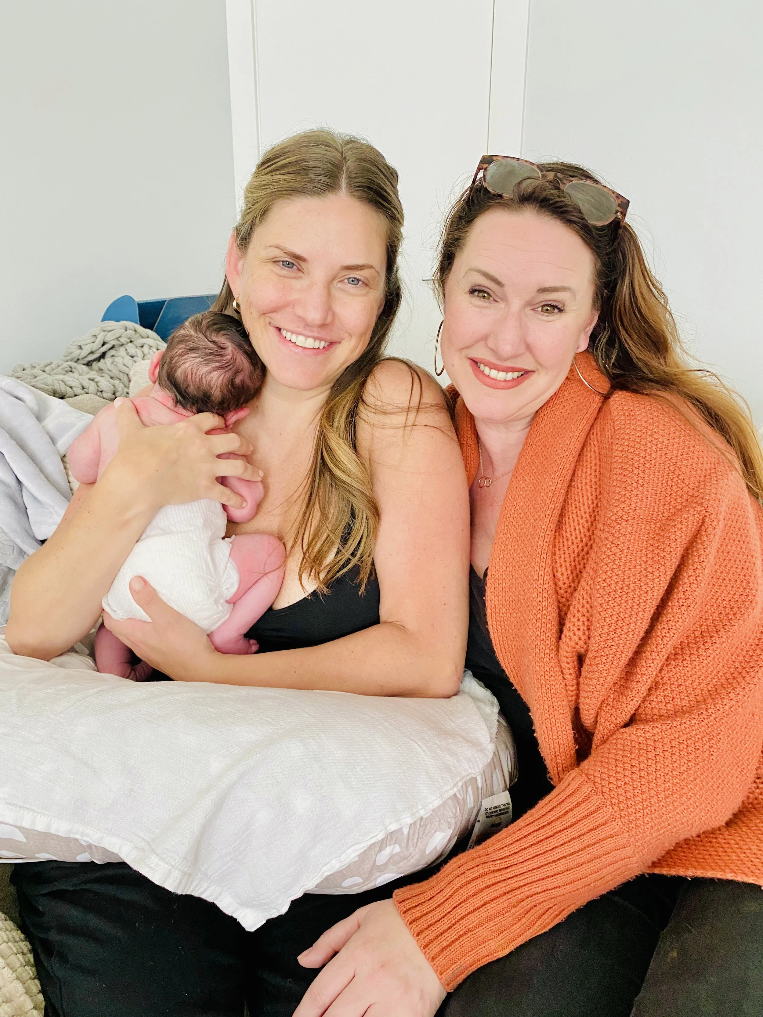 Two smiling women holding a newborn baby, with the woman on the left in a black top and the woman on the right in an orange sweater, inside a home.