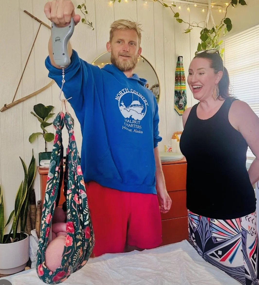 A man with a beard and a woman are smiling and looking at a scale that the man is holding up with his right hand. The man is wearing a blue hoodie and red shorts, and the woman is wearing a sleeveless black top and colorful patterned pants. 