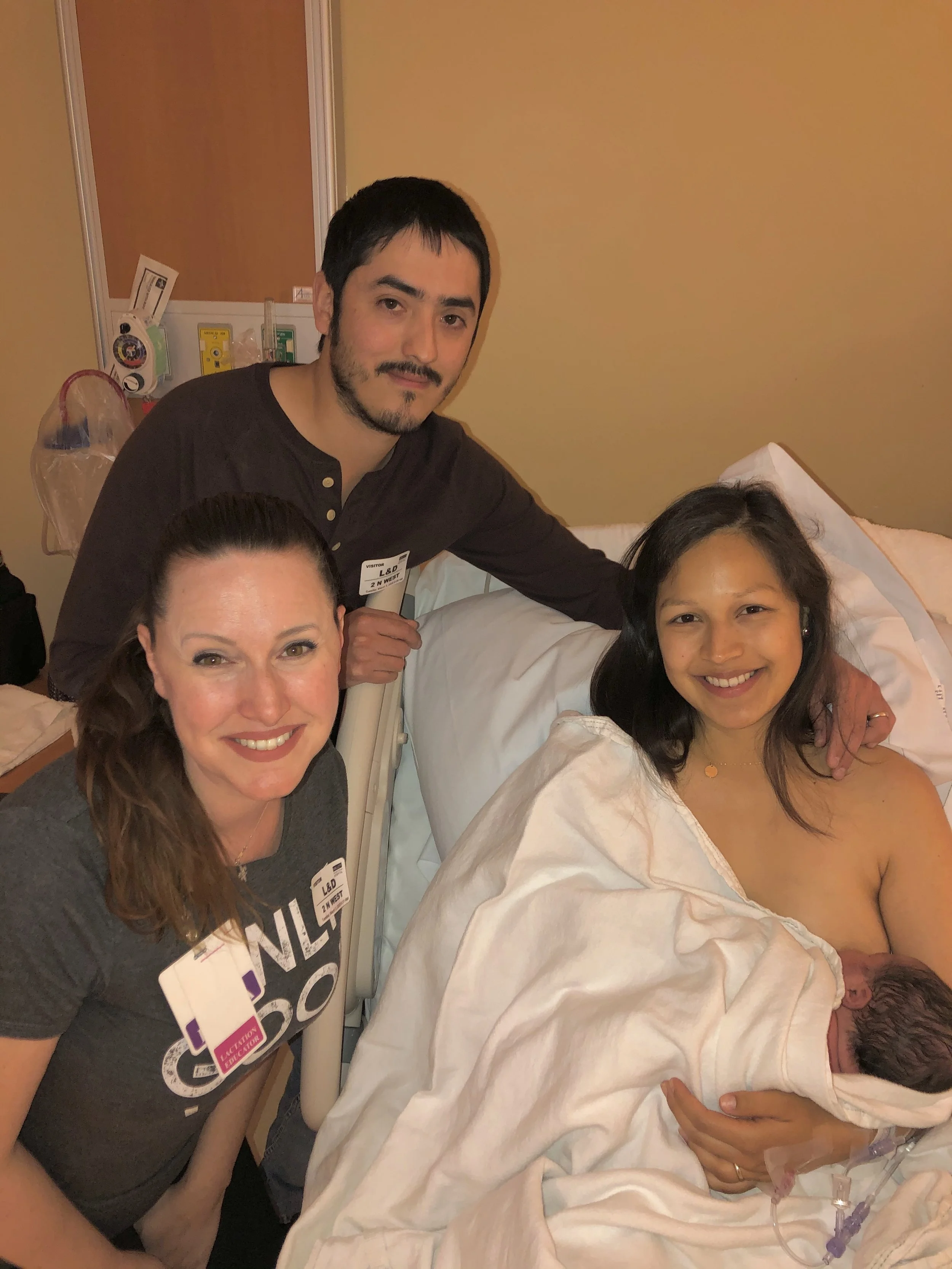 A woman who just gave birth is laying in a hospital bed holding her newborn baby, with her partner and doula surrounding her in a hospital room.