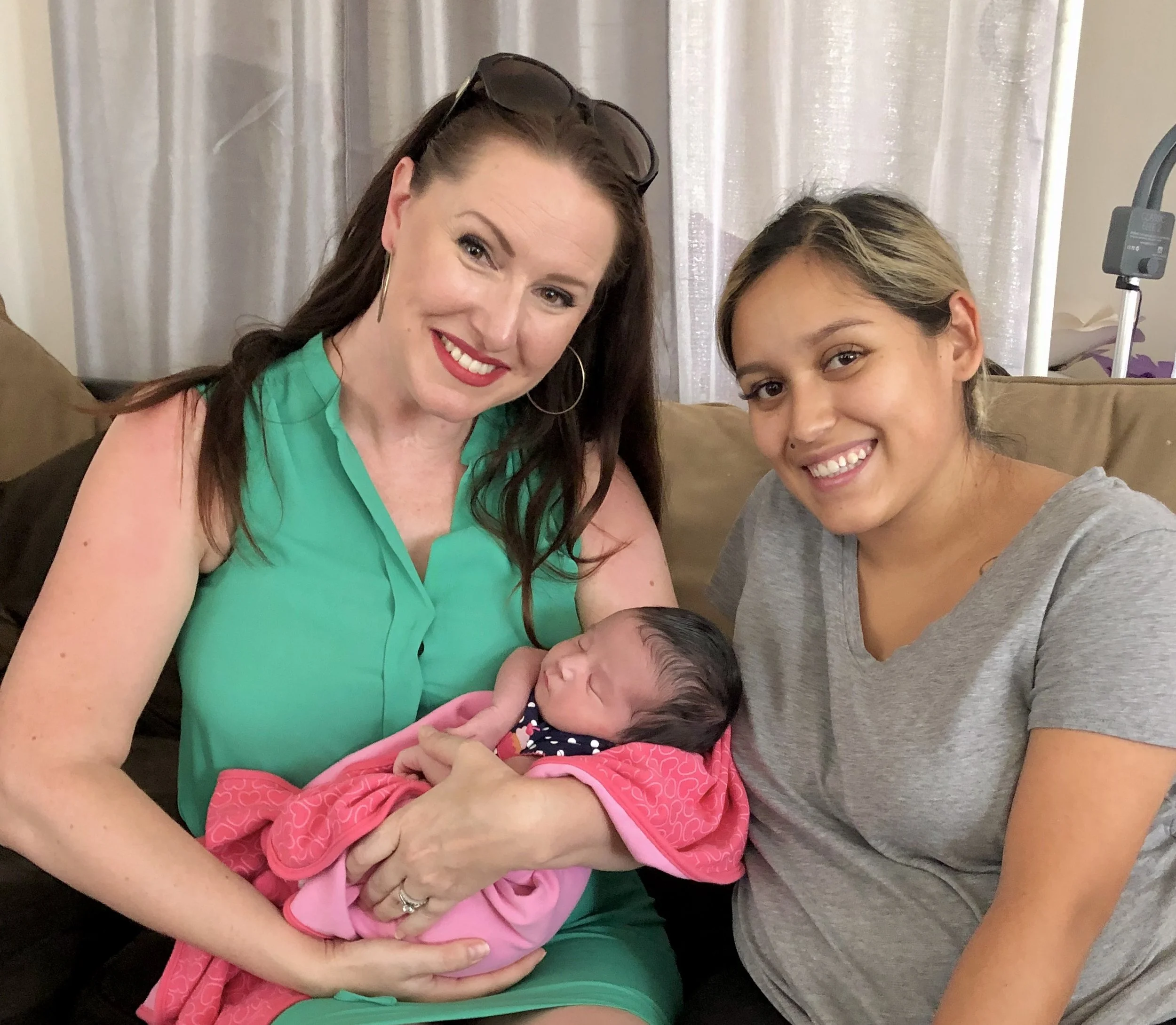 Two women sitting on a couch holding a newborn baby girl wrapped in pink blanket, smiling at the camera.