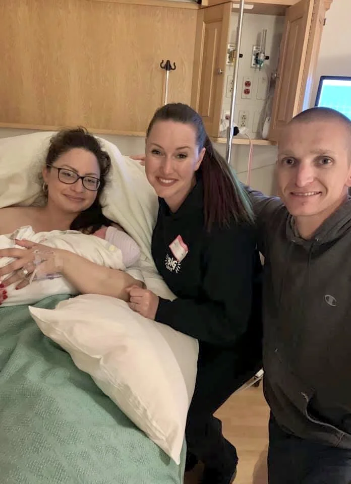 A woman in a hospital bed just gave birth and is holding hands with her doula. Her partner is standing besides them, they are all smiling. The woman in bed appears to be a patient; the others are visitors.