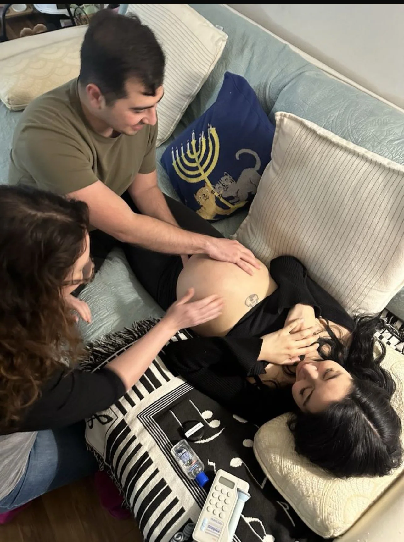 A midwife with brown hair is touching the belly of a pregnant women laying on a couch with her husband present. 