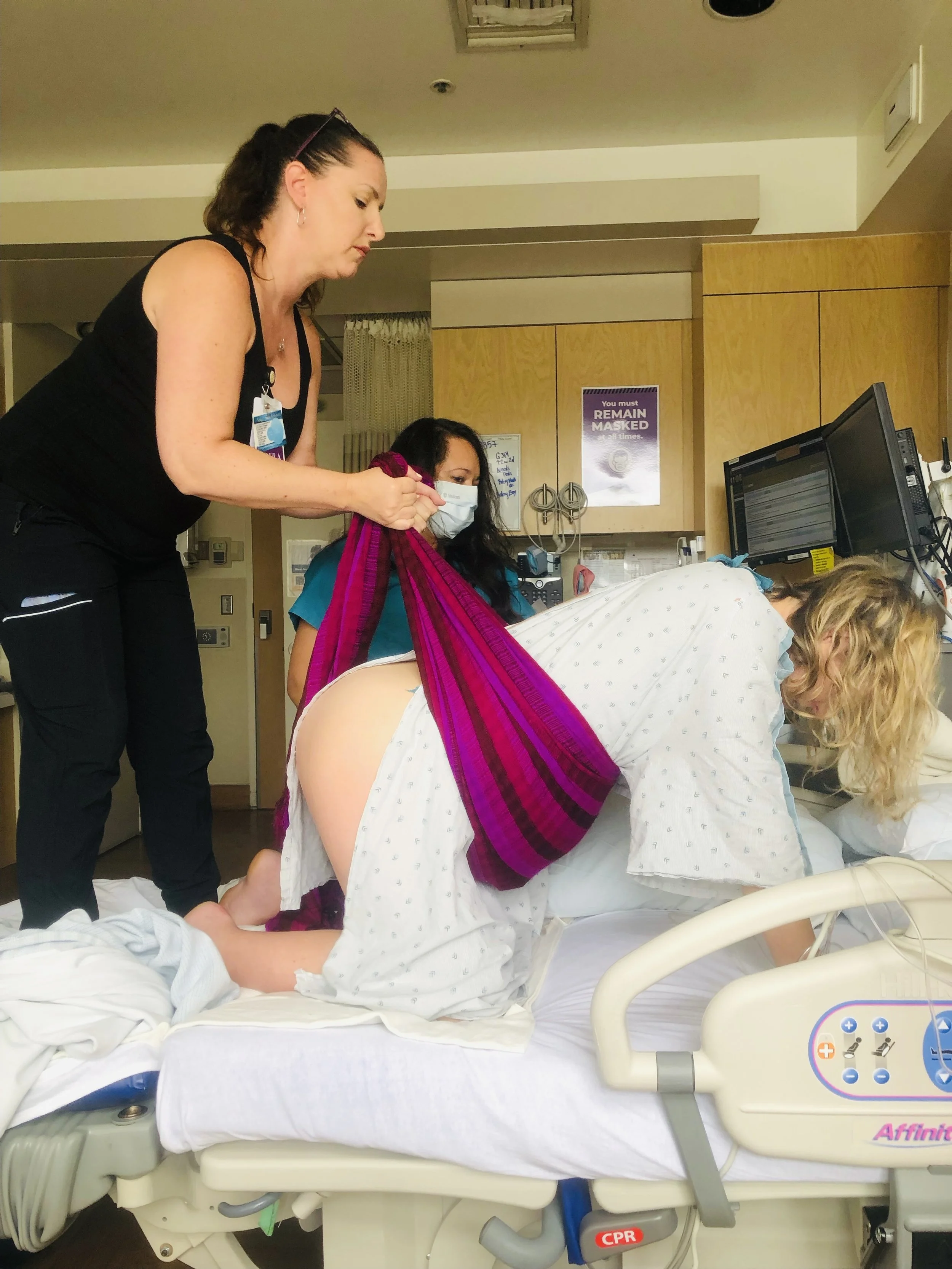 A laboring woman in a hospital bed leaning forward on all fours while a her midwife aids her.