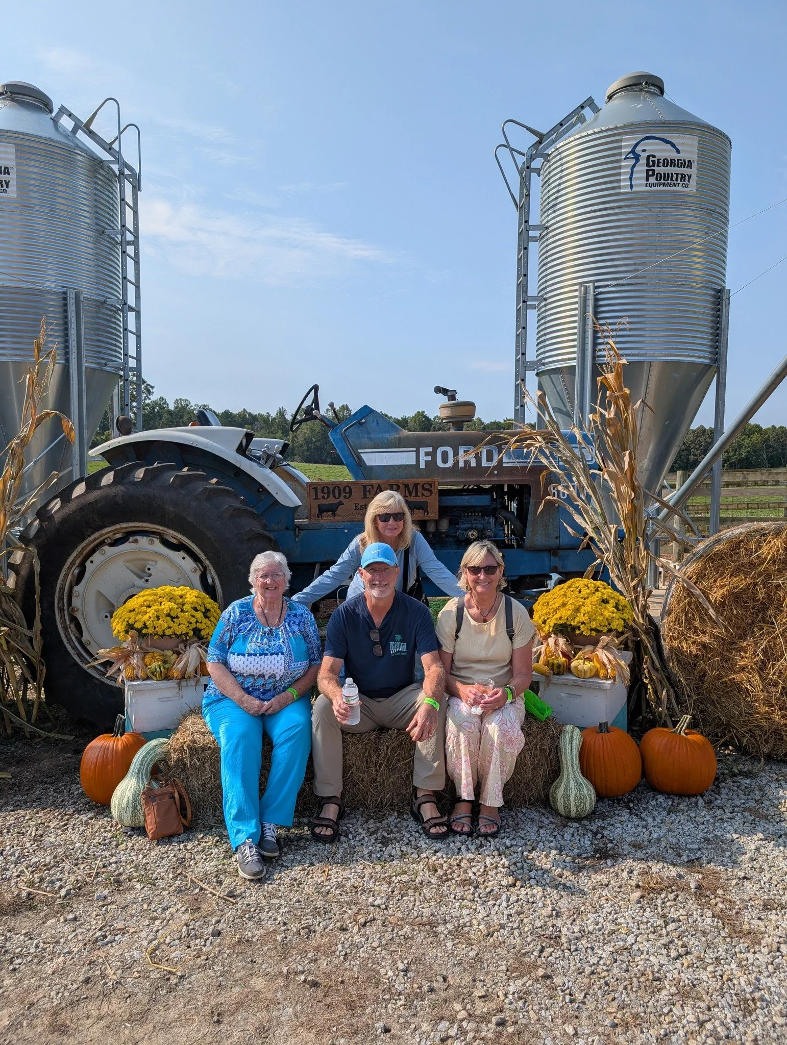 Four people sitting on hay bales in front of a vintage blue tractor with pumpkins, gourds, and yellow chrysanthemums, surrounded by metal grain silos, outdoors on a farm.