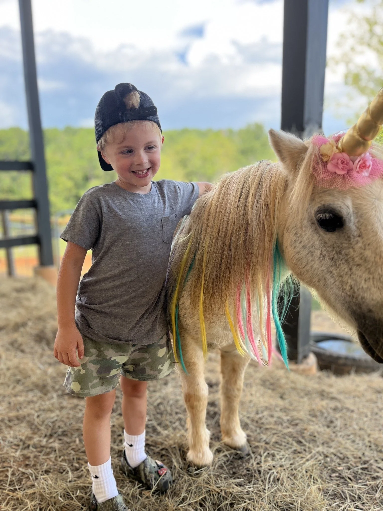 A young boy wearing a gray t-shirt, camouflage shorts, and a backwards black baseball cap, standing next to a small pony with a rainbow-colored mane and a golden unicorn horn, on a farm or petting zoo setting with trees in the background.
