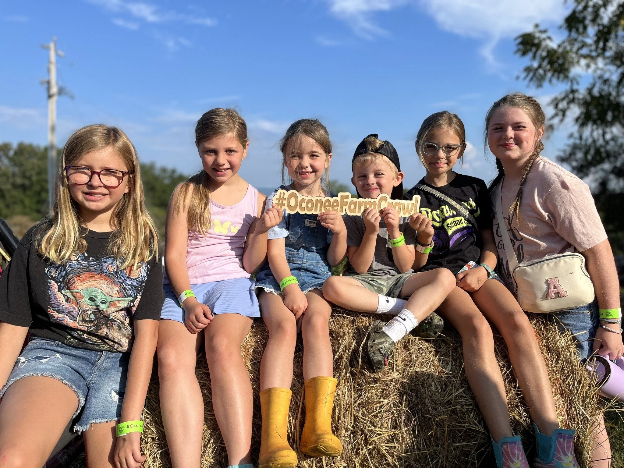 Group of six children sitting on hay bales outdoors on a sunny day, holding a hashtag sign that says '#OconeeFarmsCrawl', smiling at the camera.