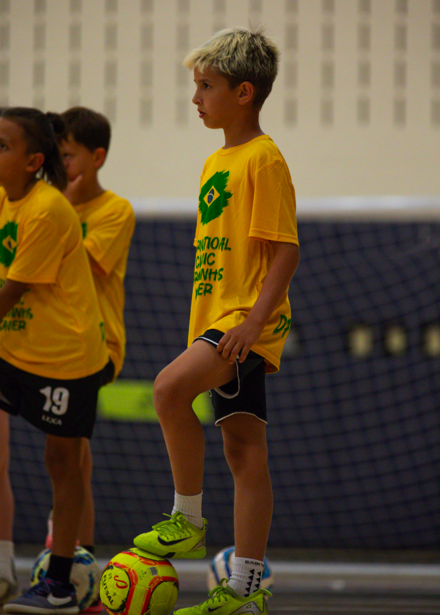A young boy in yellow sportswear with a Brazil flag emblem and text, standing on a football in an indoor gym, balancing with one foot on the ball while others stand nearby.