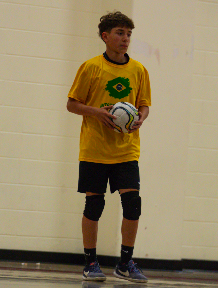 A young male athlete wearing a yellow Brazil soccer jersey, black shorts, and knee pads, holding a soccer ball indoors.