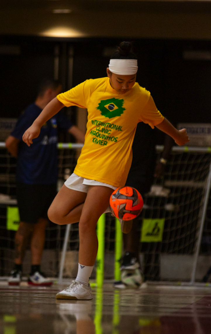 A young girl in a yellow shirt and white shorts is playing indoor soccer with an orange and gray ball. She is wearing a white headband and white sneakers, focusing on kicking the ball. In the background, there are other children and a net, indicating a sports facility.