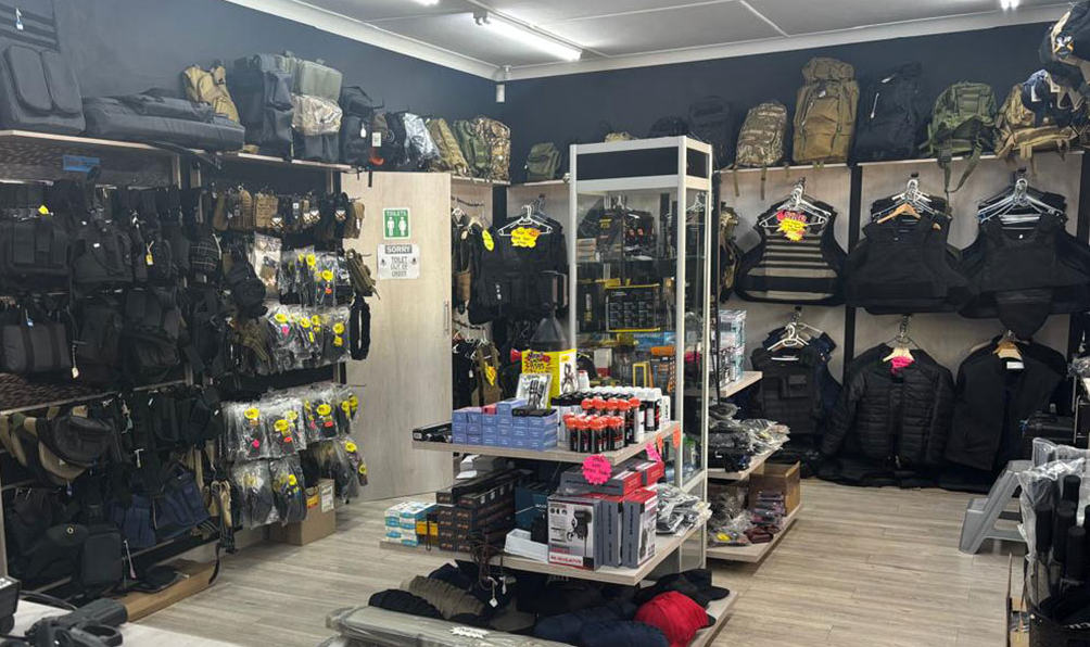 Inne i en butik med militär- eller outdoor-utrustning, med väskor, ryggsäckar, kläder och tillbehör på hyllor och väggar.