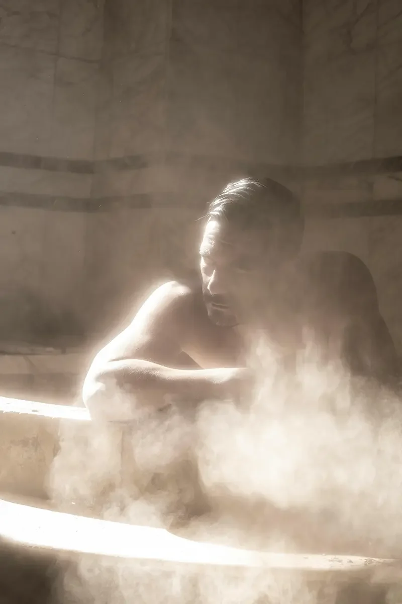 A man with short dark hair and facial hair, shirtless, relaxing in a steam room with stone walls, illuminated by soft light.