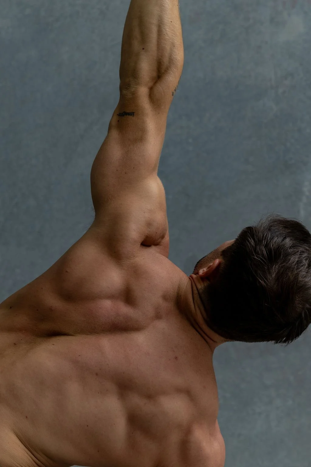 A shirtless man with dark hair performing a stretch or yoga pose, with his arm raised above his head and his body tilted to the side against a plain, textured gray background.