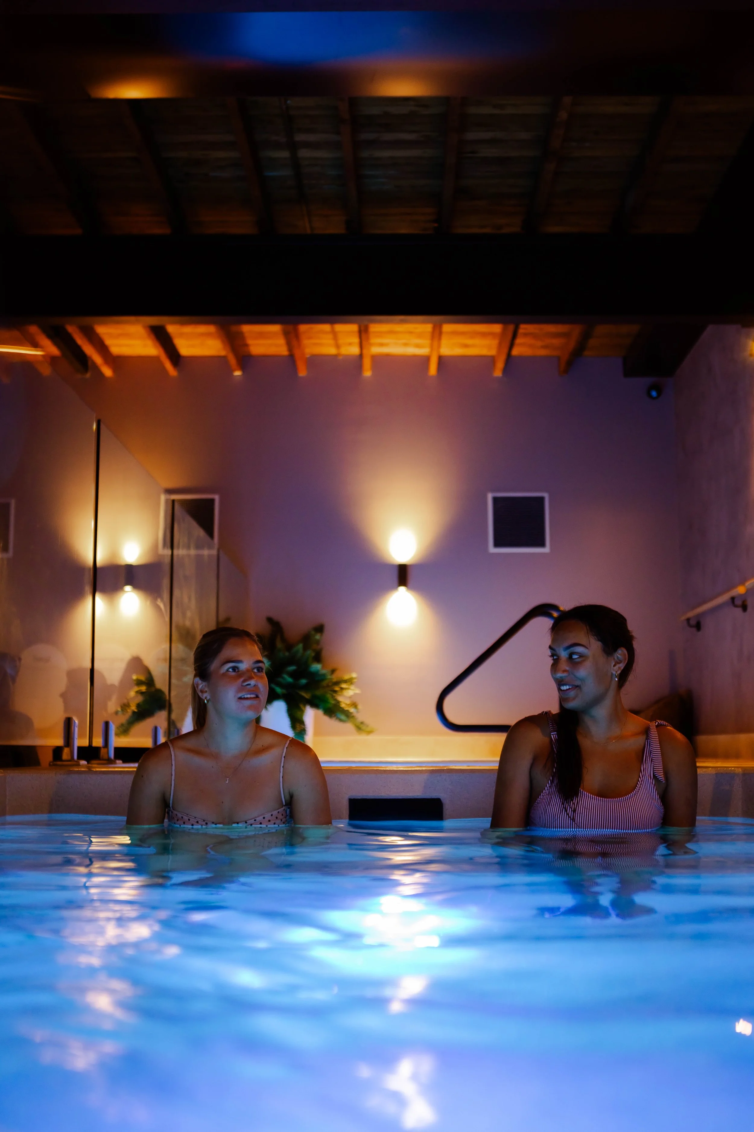 Two girls sitting in a cold plunge, looking at one another while doing a bathhouse session.